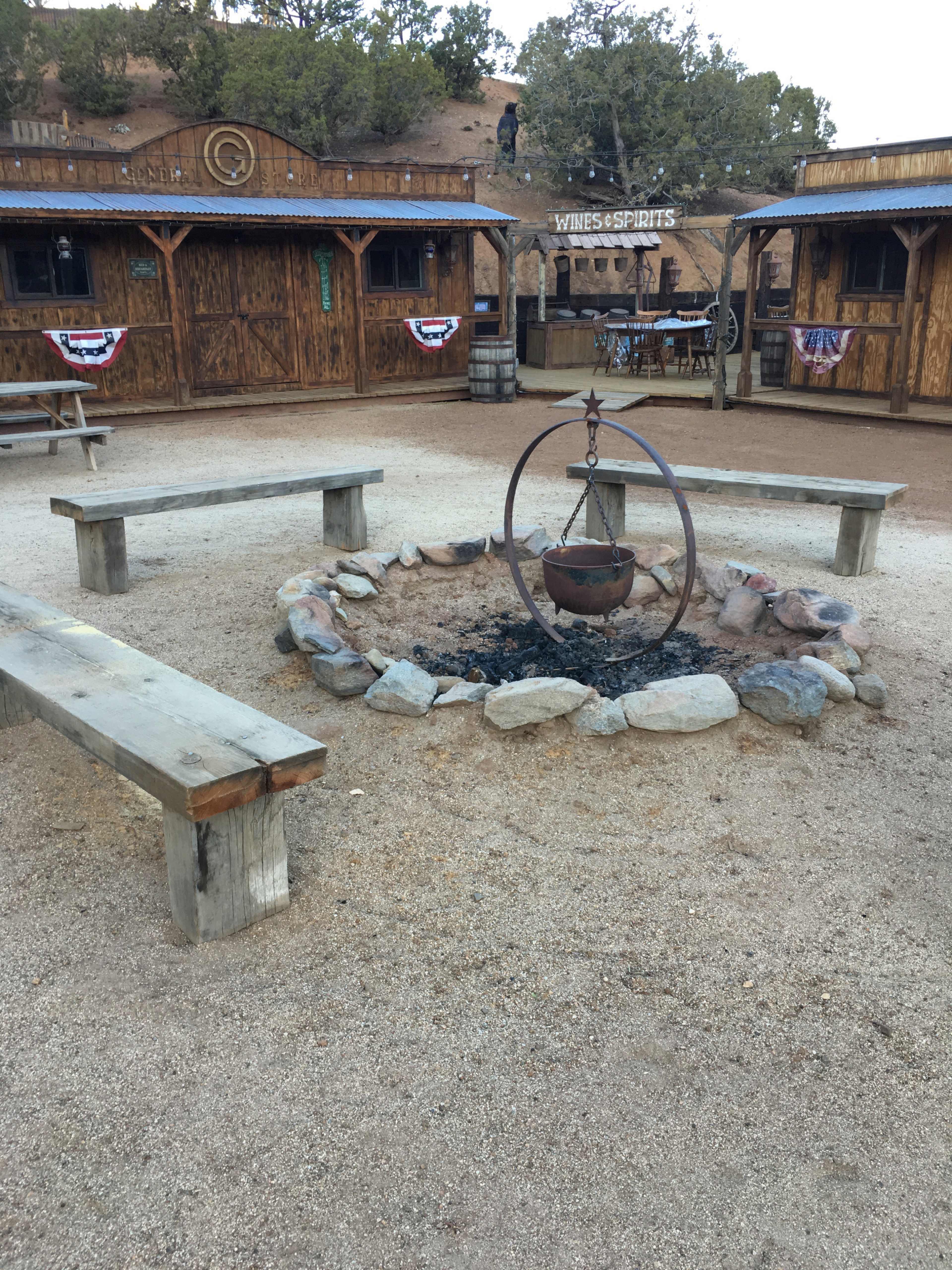 A circular fire pit surrounded by wooden benches in a rustic outdoor setting with wooden structures in the background.