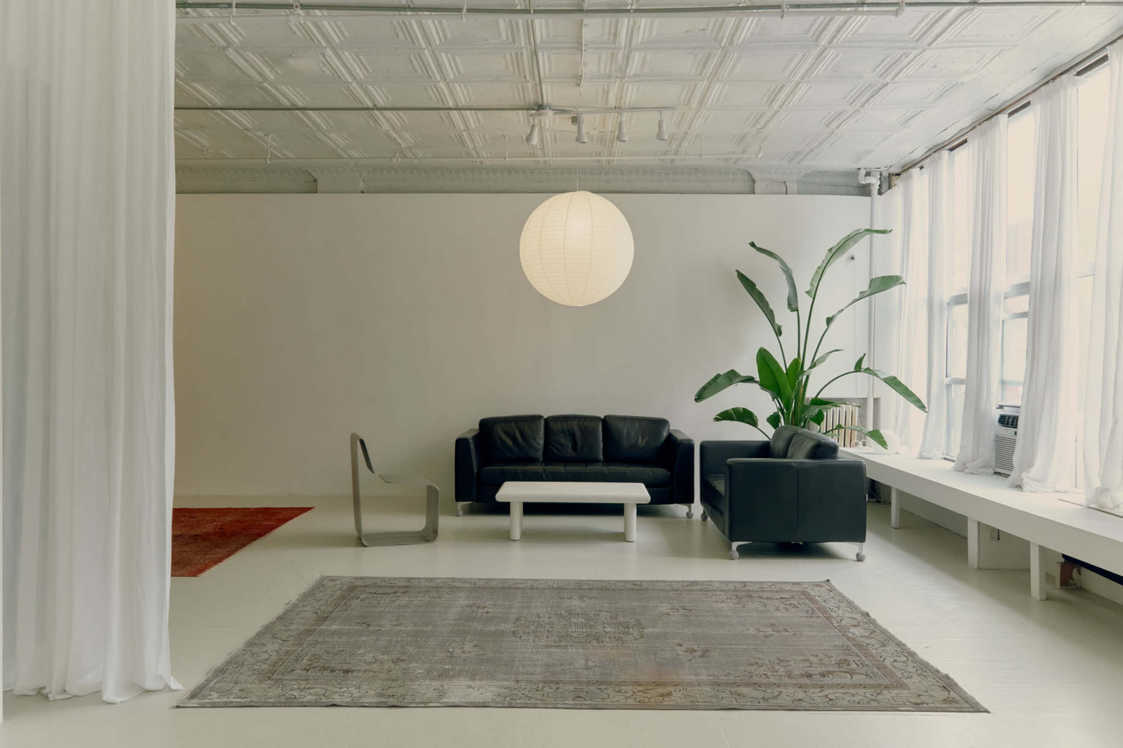 The image shows a minimalistic living space featuring a black sofa set, a large plant, a round paper lantern, and a decorative rug on a light-colored floor.