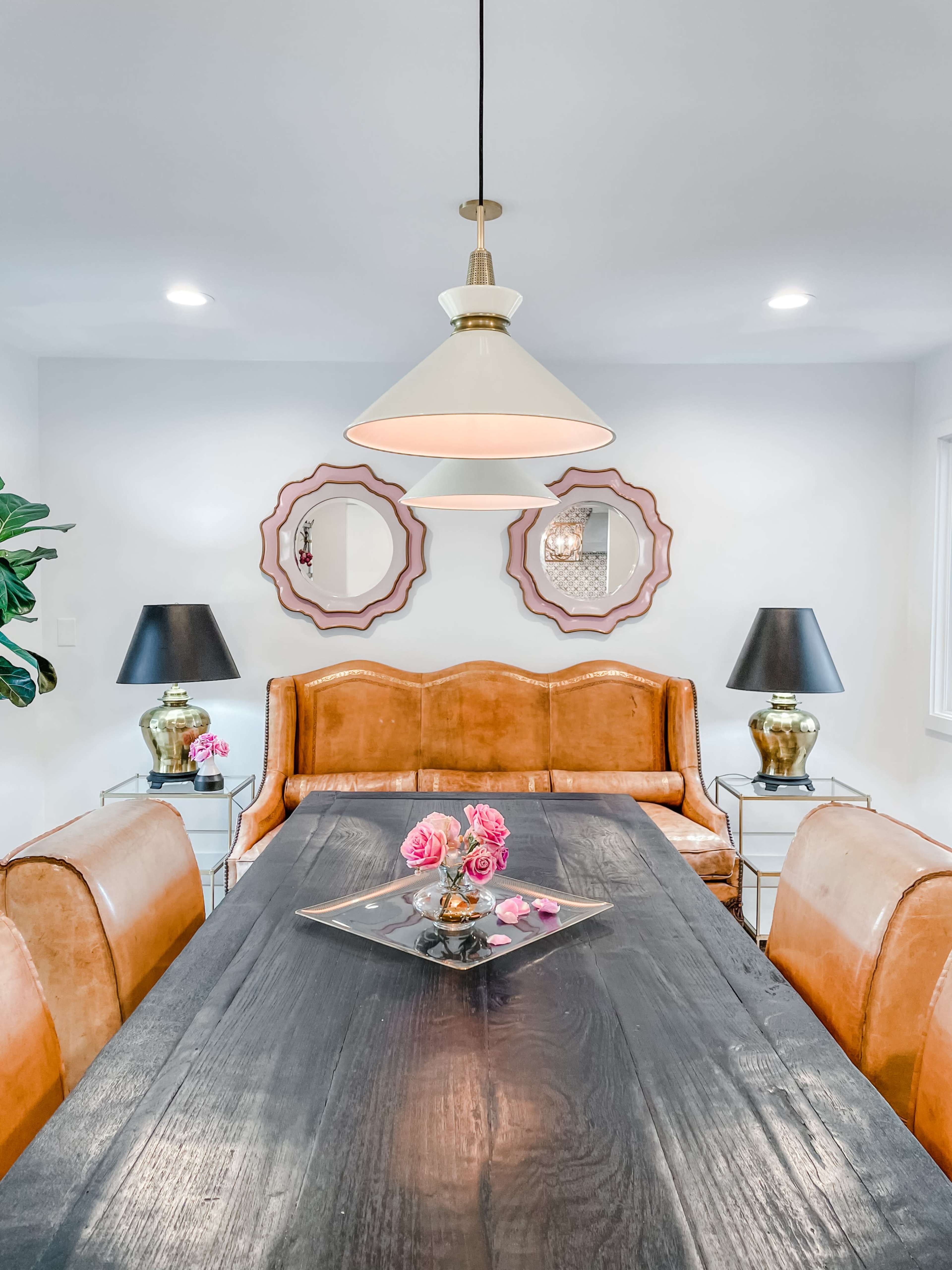 A dining area featuring a brown leather sofa, a dark wood dining table, and decorative lamps, with pink flowers as a centerpiece.