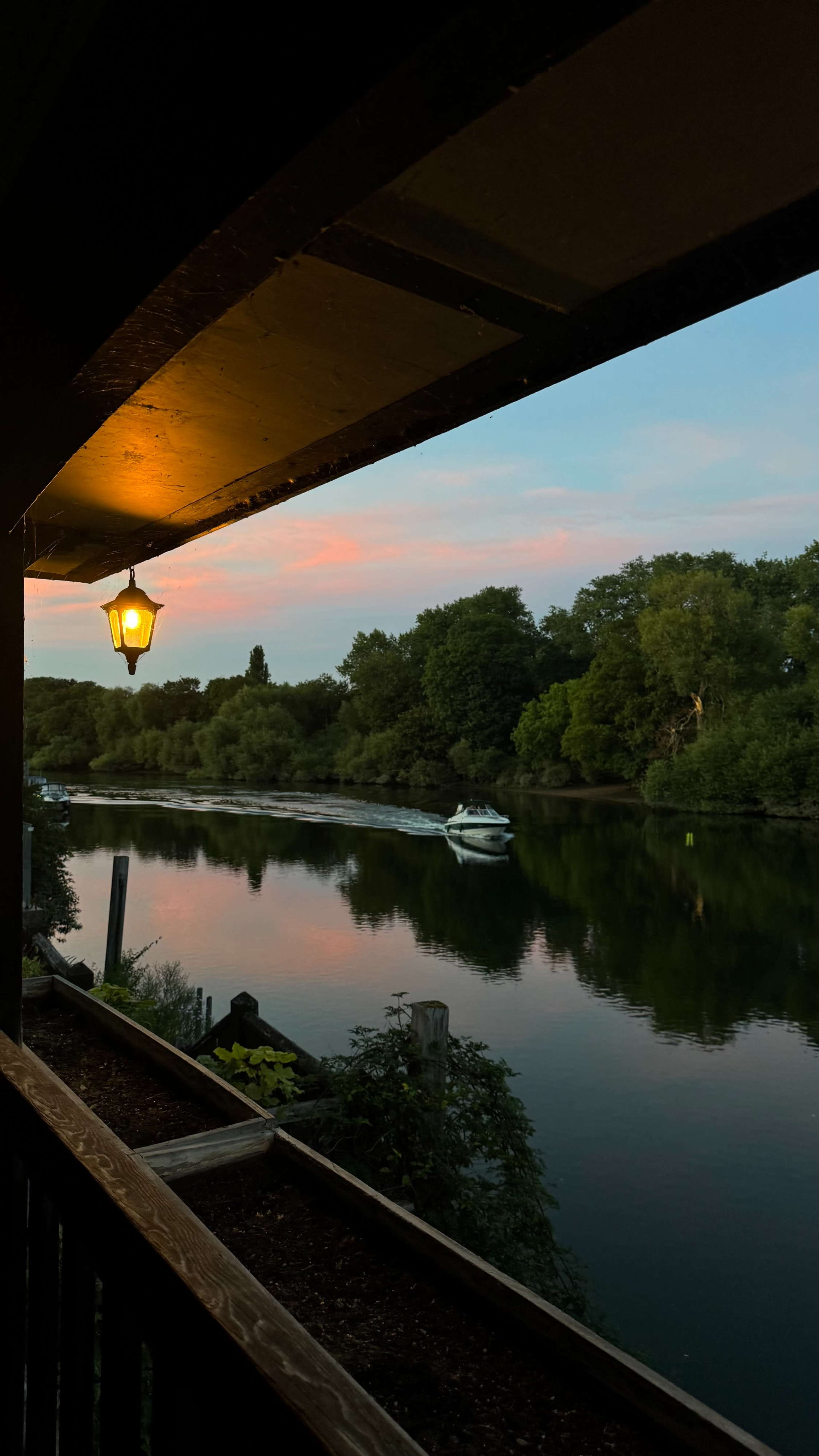 The image shows a calm river with a small boat drifting past, framed by a wooden structure and lush greenery under a colorful sky at dusk.
