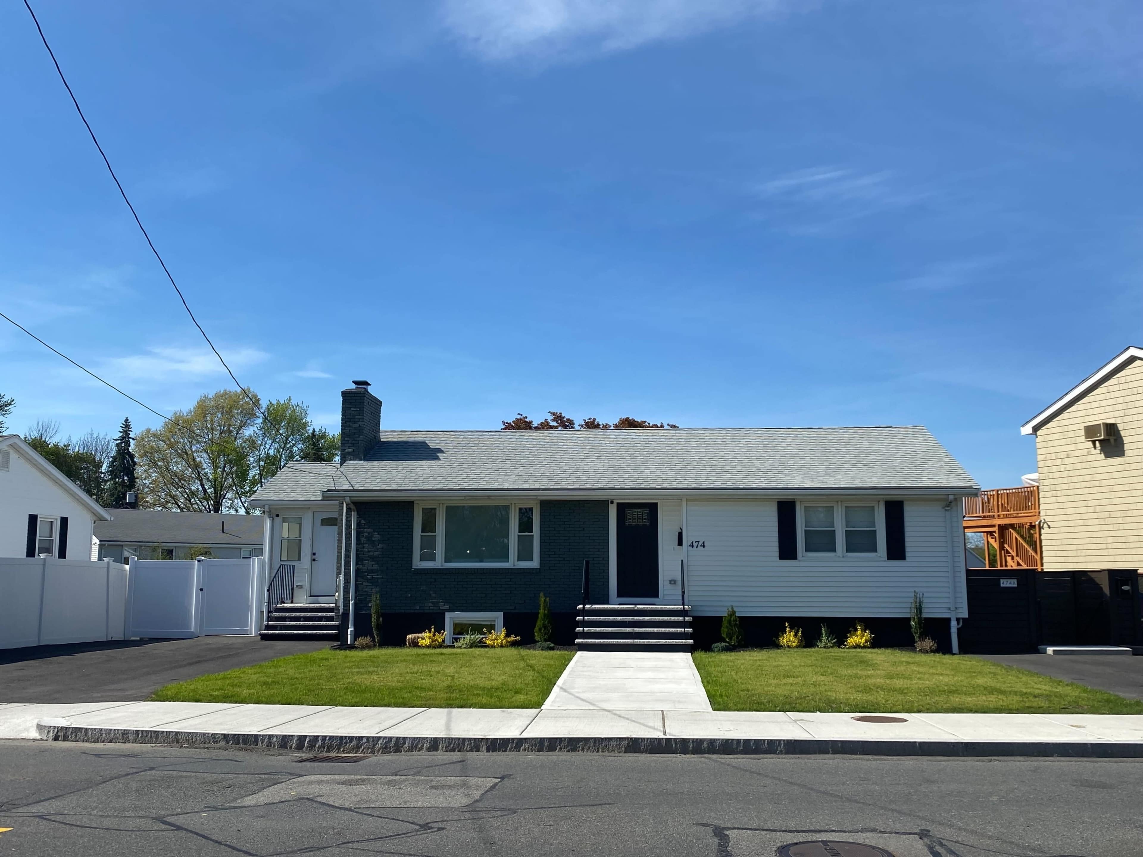 A single-story house with gray siding and a well-manicured lawn is situated at the corner of a residential street under a clear blue sky.