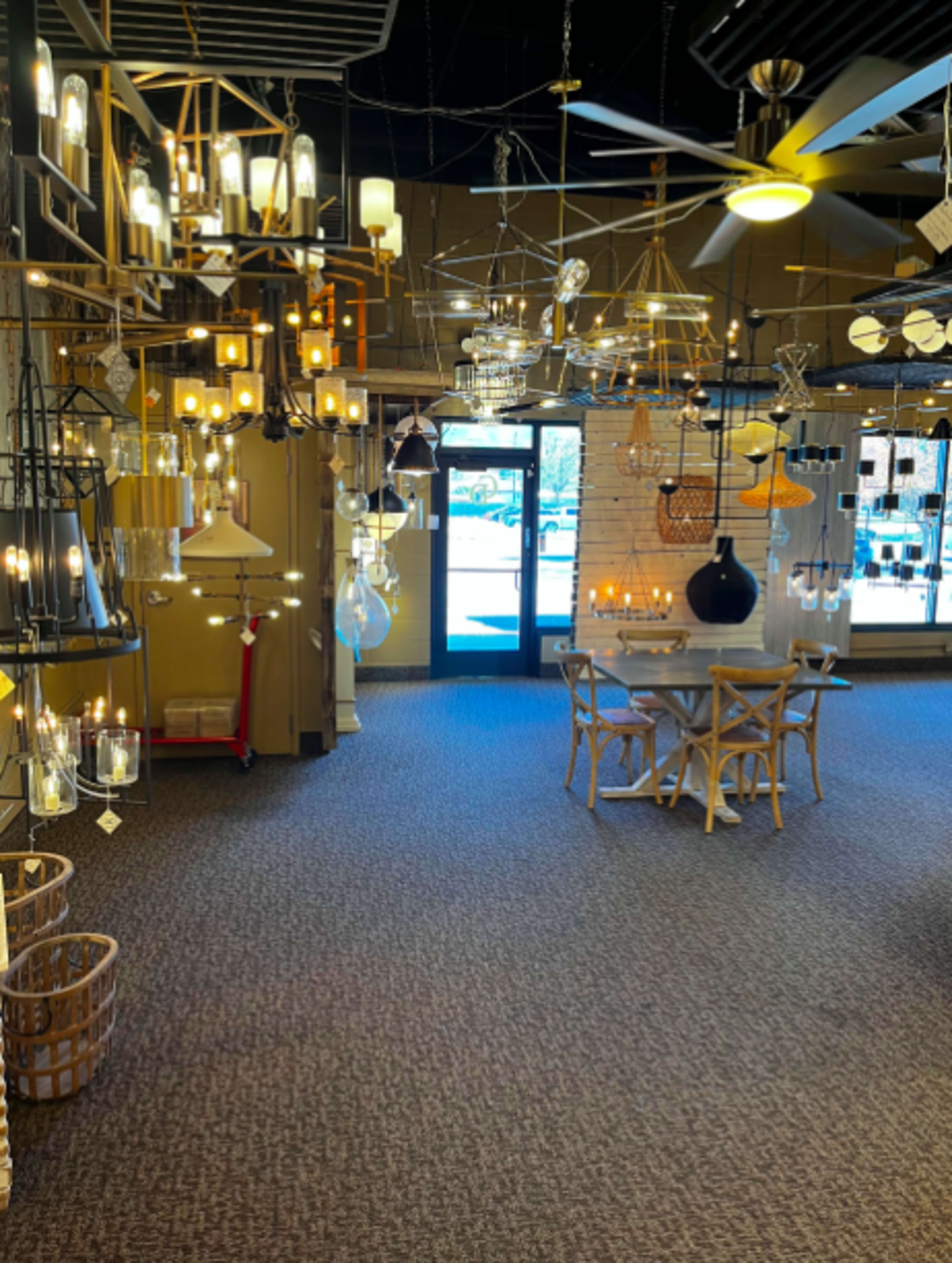 A well-lit showroom displaying various hanging light fixtures with a dining table and chairs in the center.