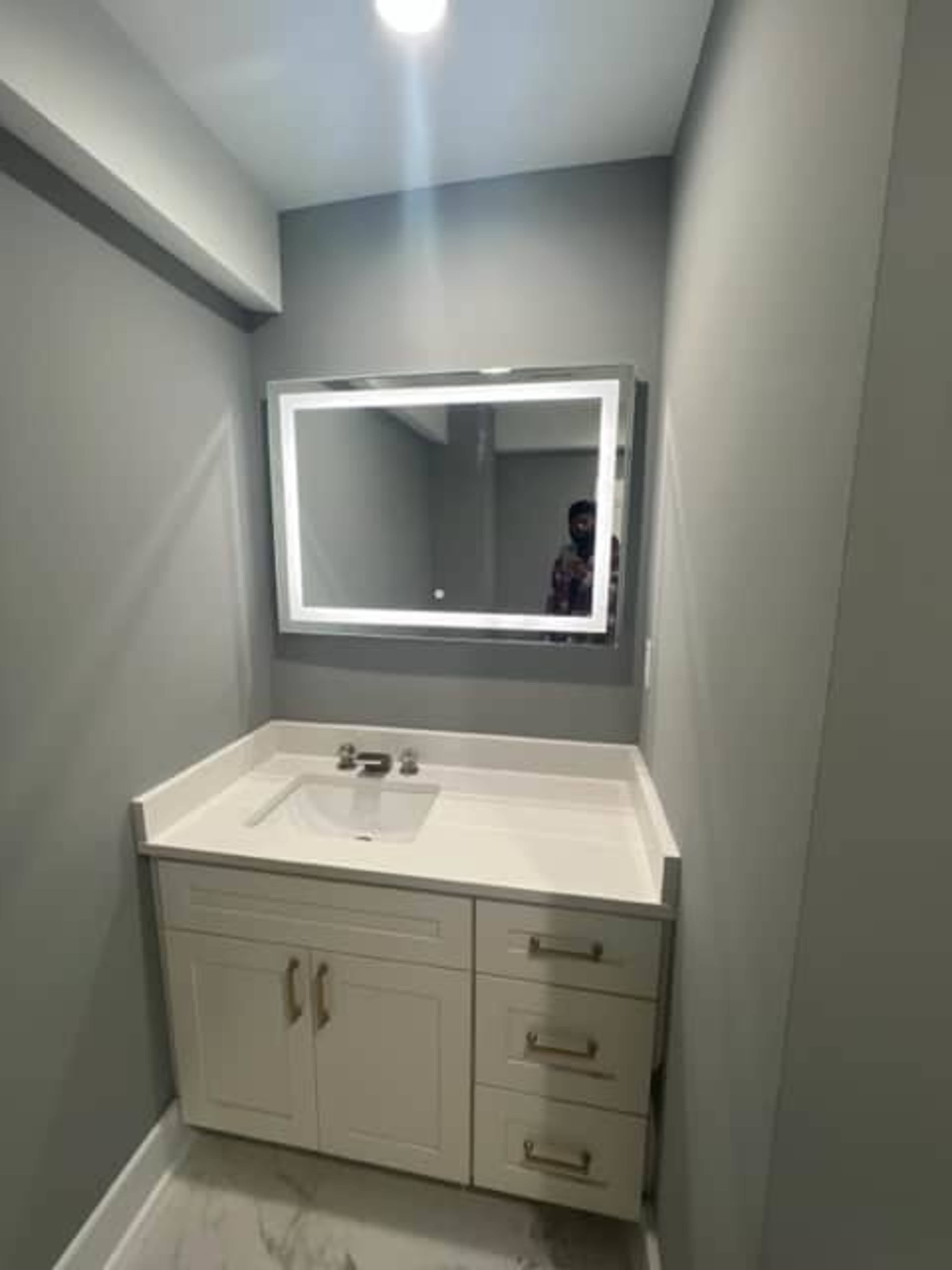 The image shows a modern bathroom with a white sink and cabinetry beneath a backlit mirror against gray walls.