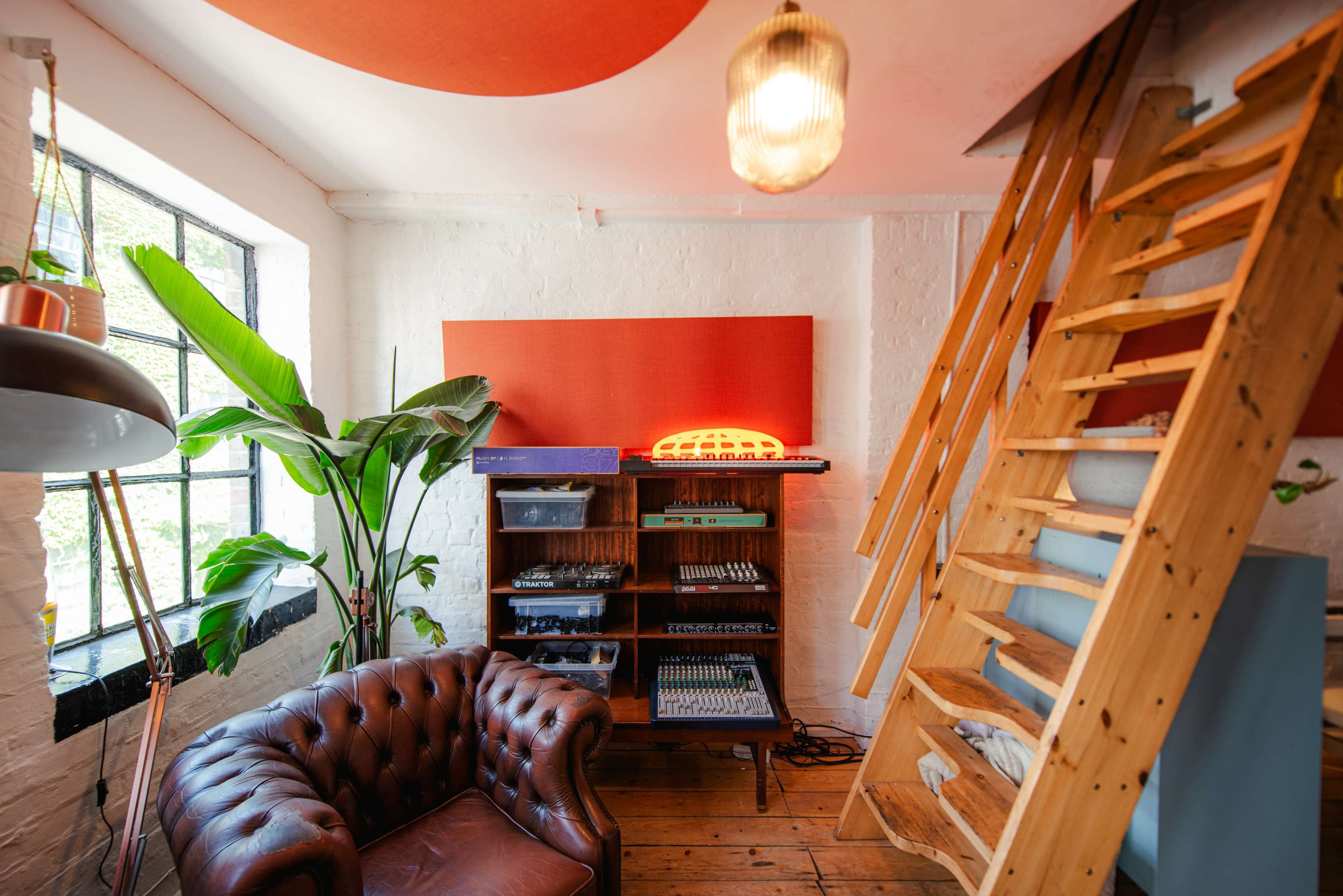 The image shows a cozy room with a leather couch, wooden stairs, a shelving unit with various items, and a large window with a leafy plant nearby.