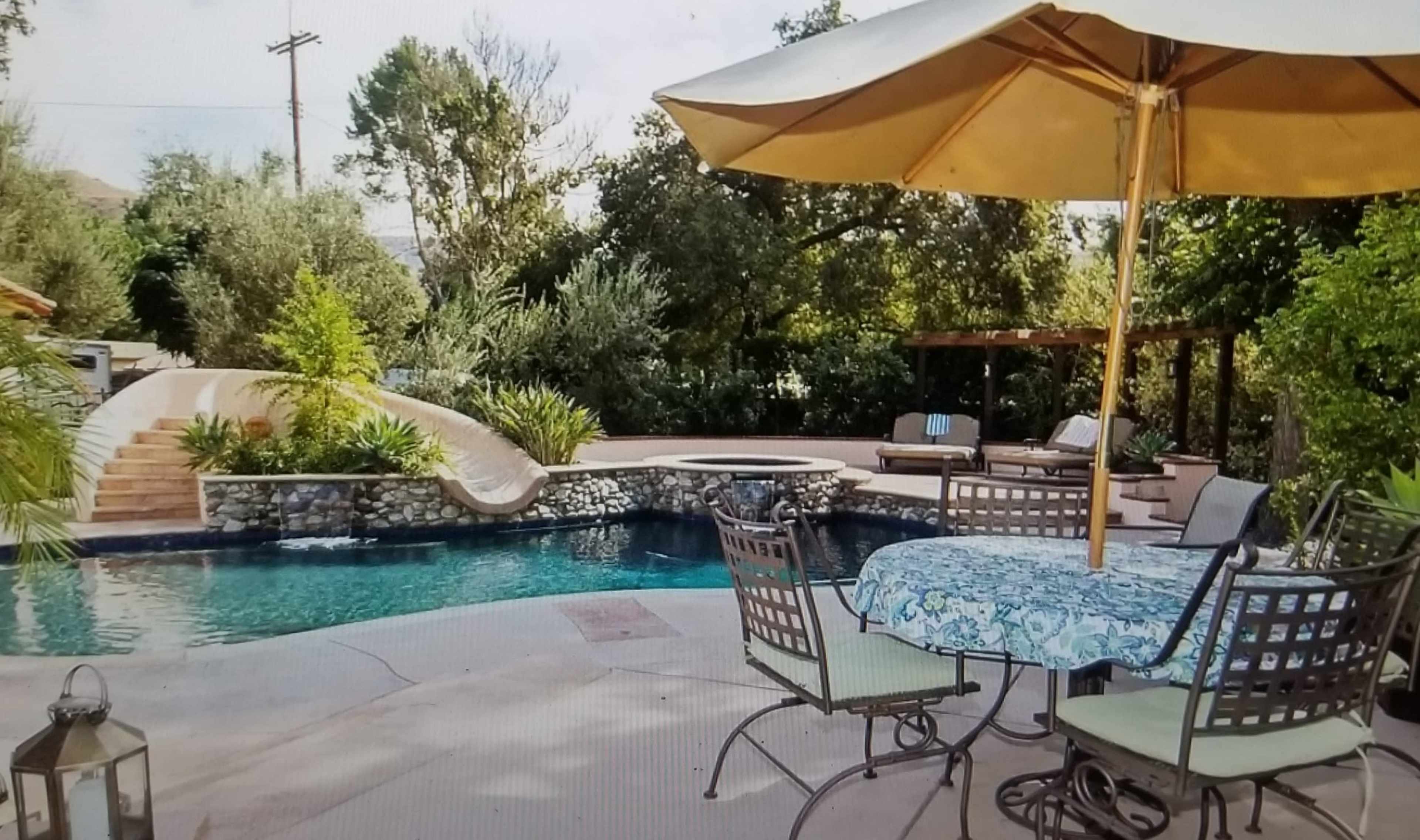 A backyard with a swimming pool, a patio table with chairs, and a large umbrella, surrounded by greenery and a stone wall.