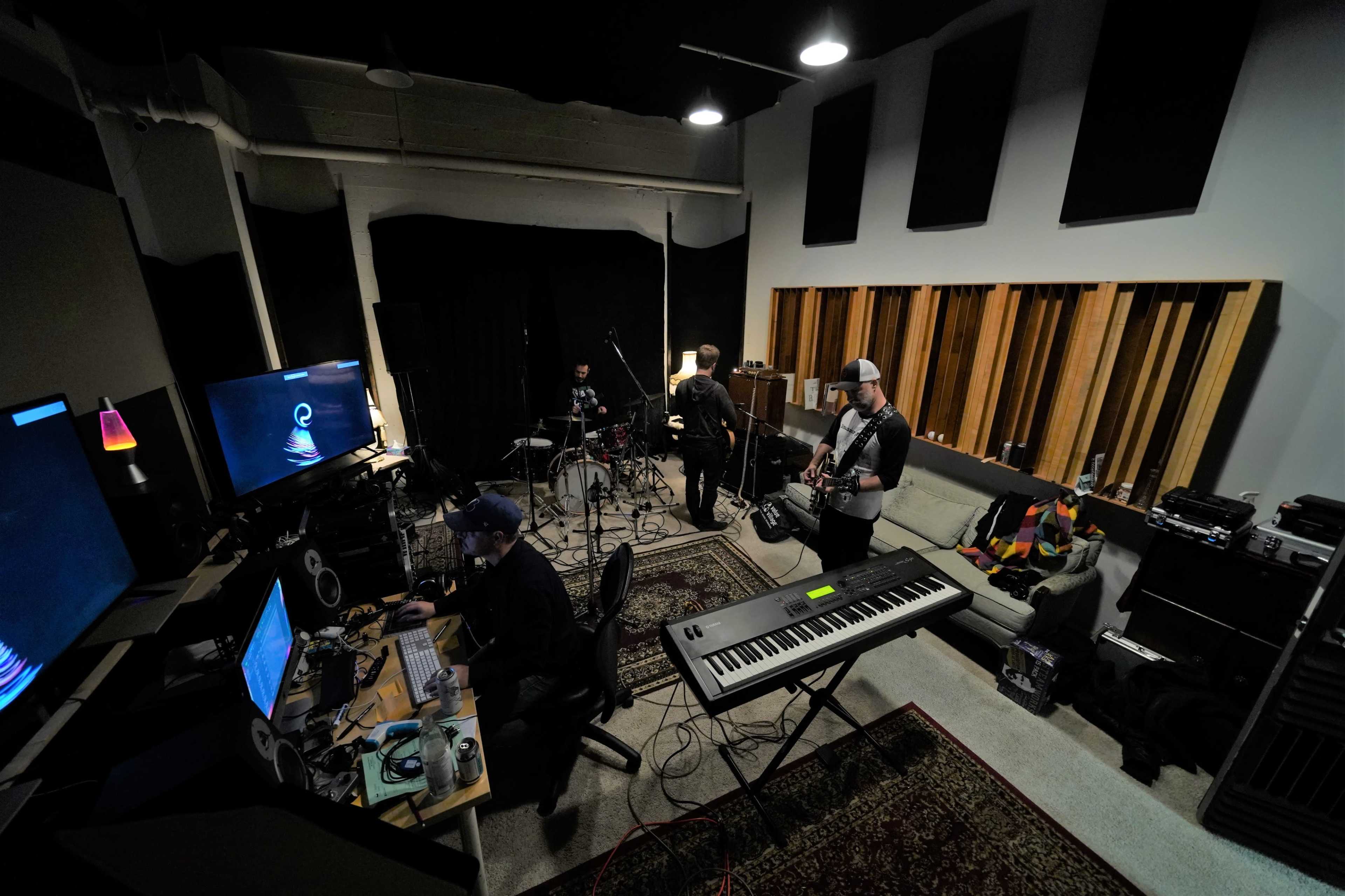 A group of musicians collaborates in a studio, with one on the keyboard, another on drums, and a guitarist in the background, surrounded by various audio equipment.