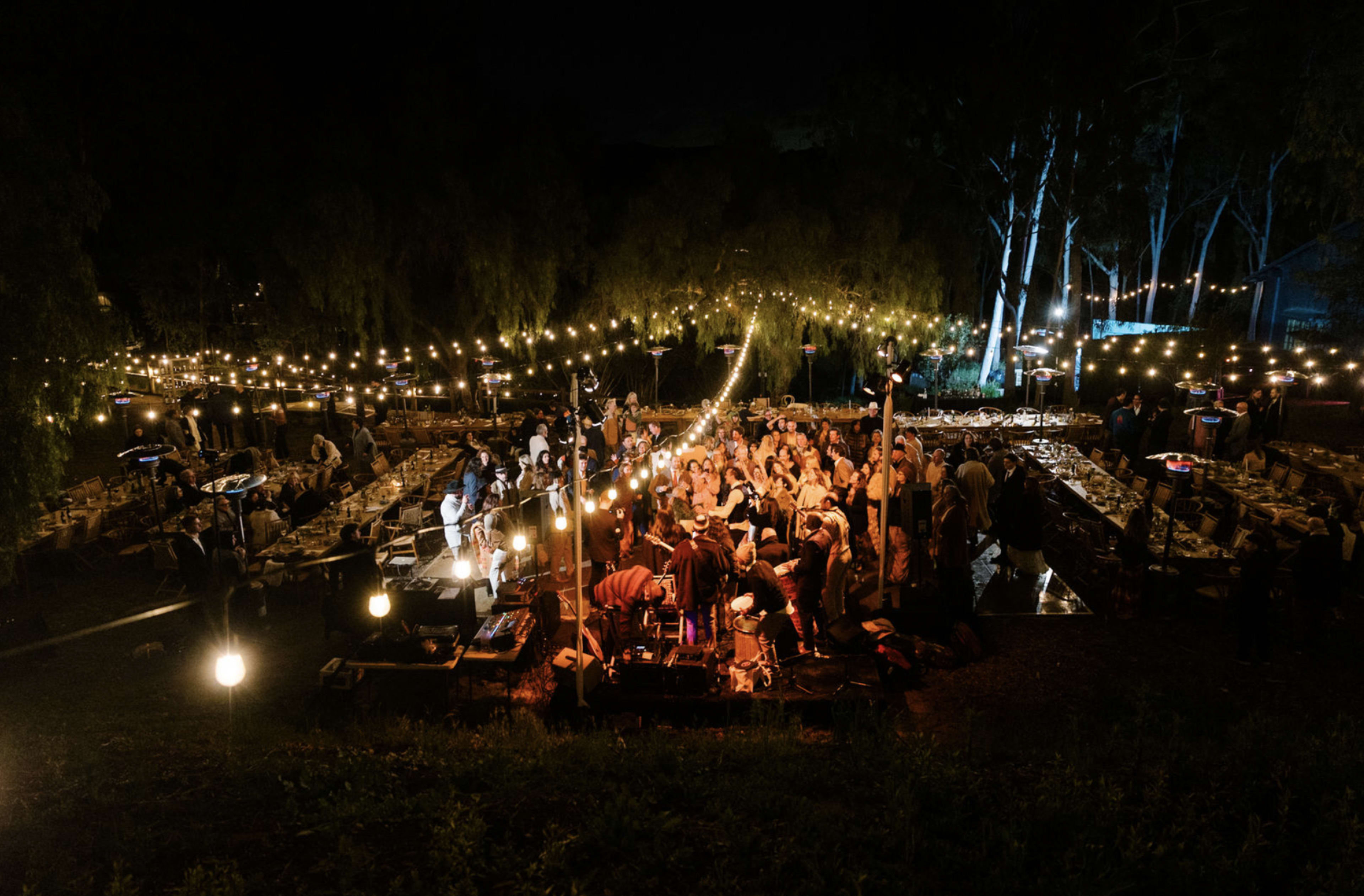 A lively outdoor gathering features numerous tables set for a feast, illuminated by string lights and surrounded by trees at night.