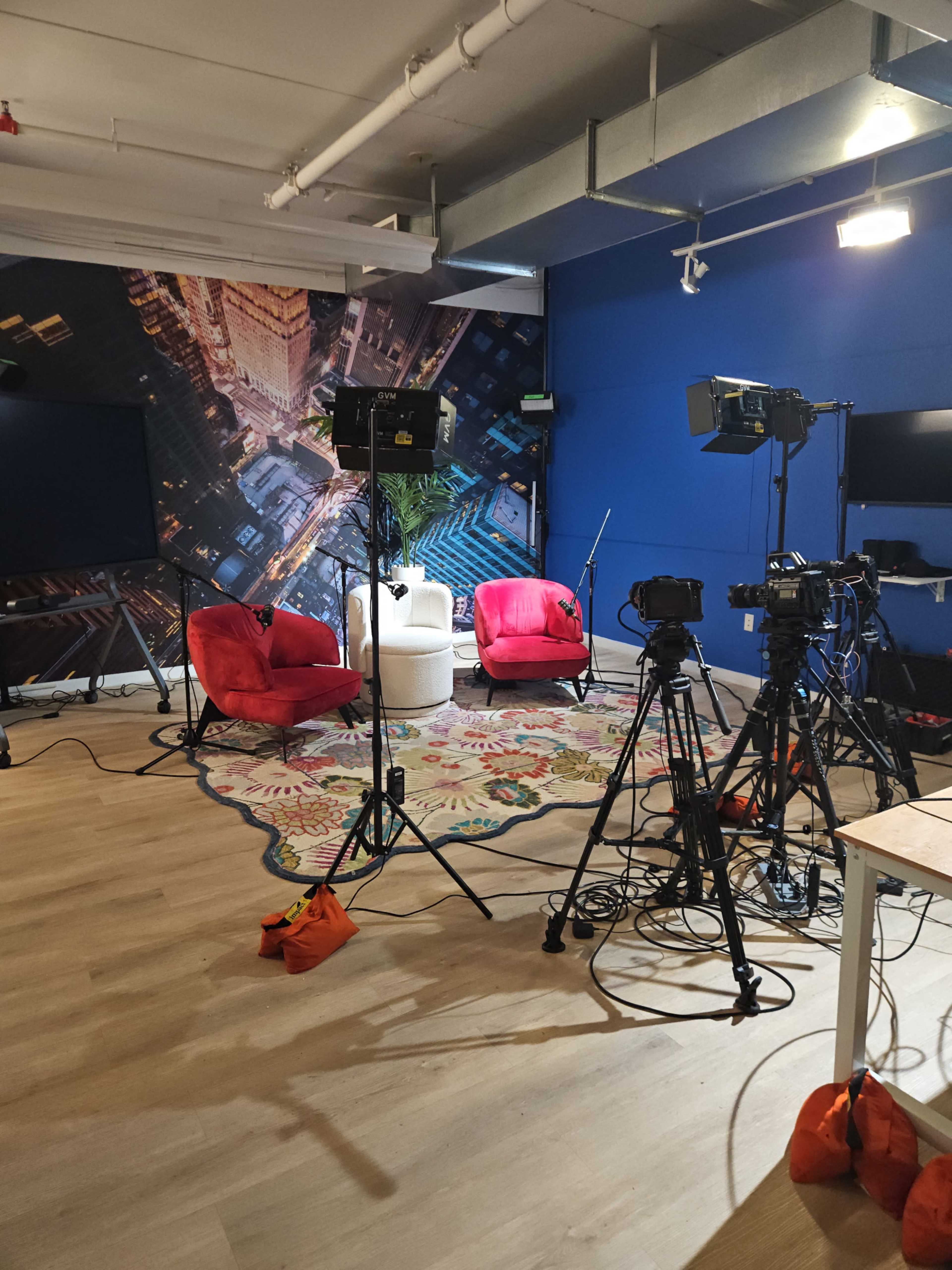 The image shows a studio setup with red chairs, multiple camera tripods, and lighting equipment, arranged in a space featuring a large urban-themed backdrop.