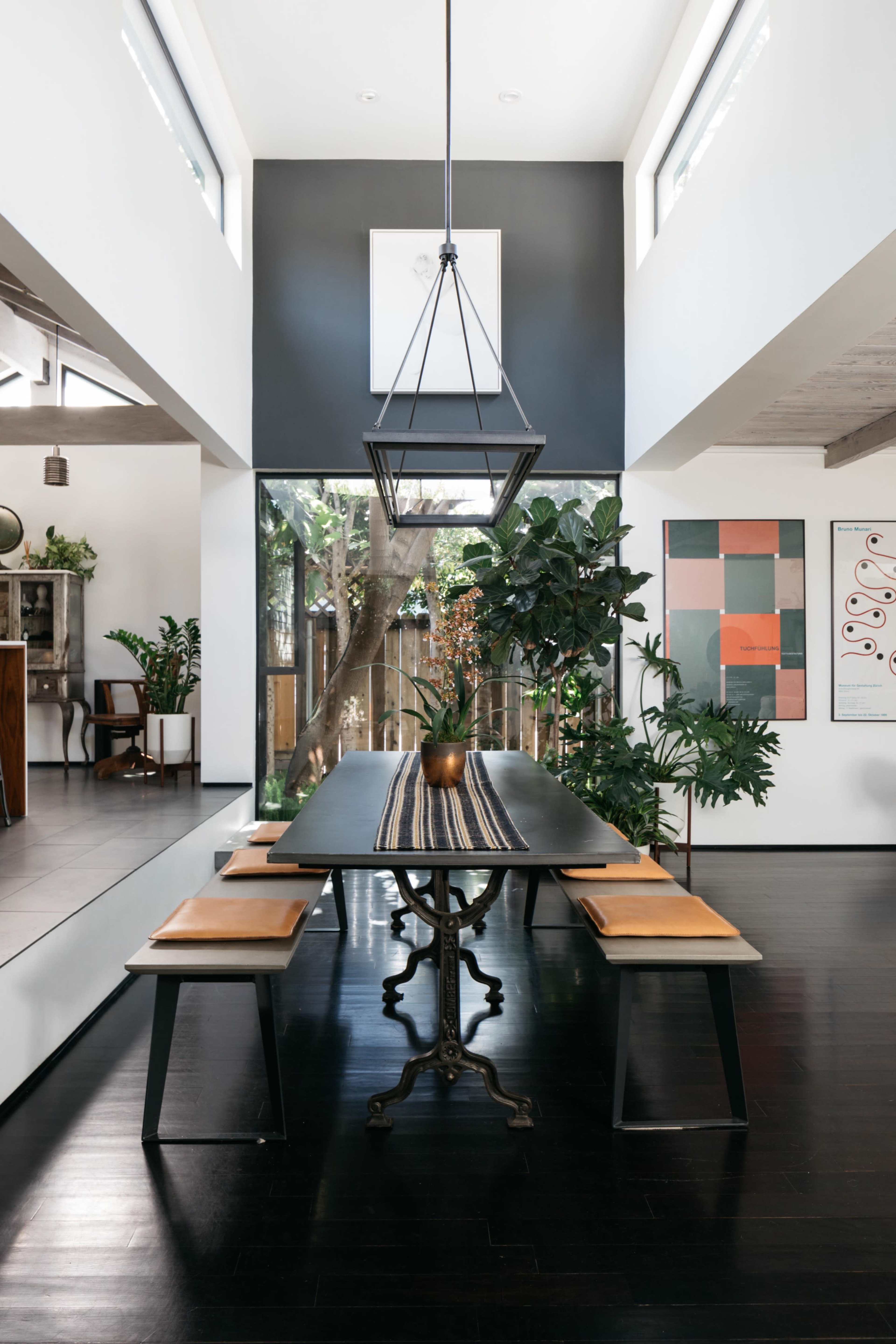 A long dining table with leather benches is set in a modern, open-concept room featuring lush greenery and large windows.