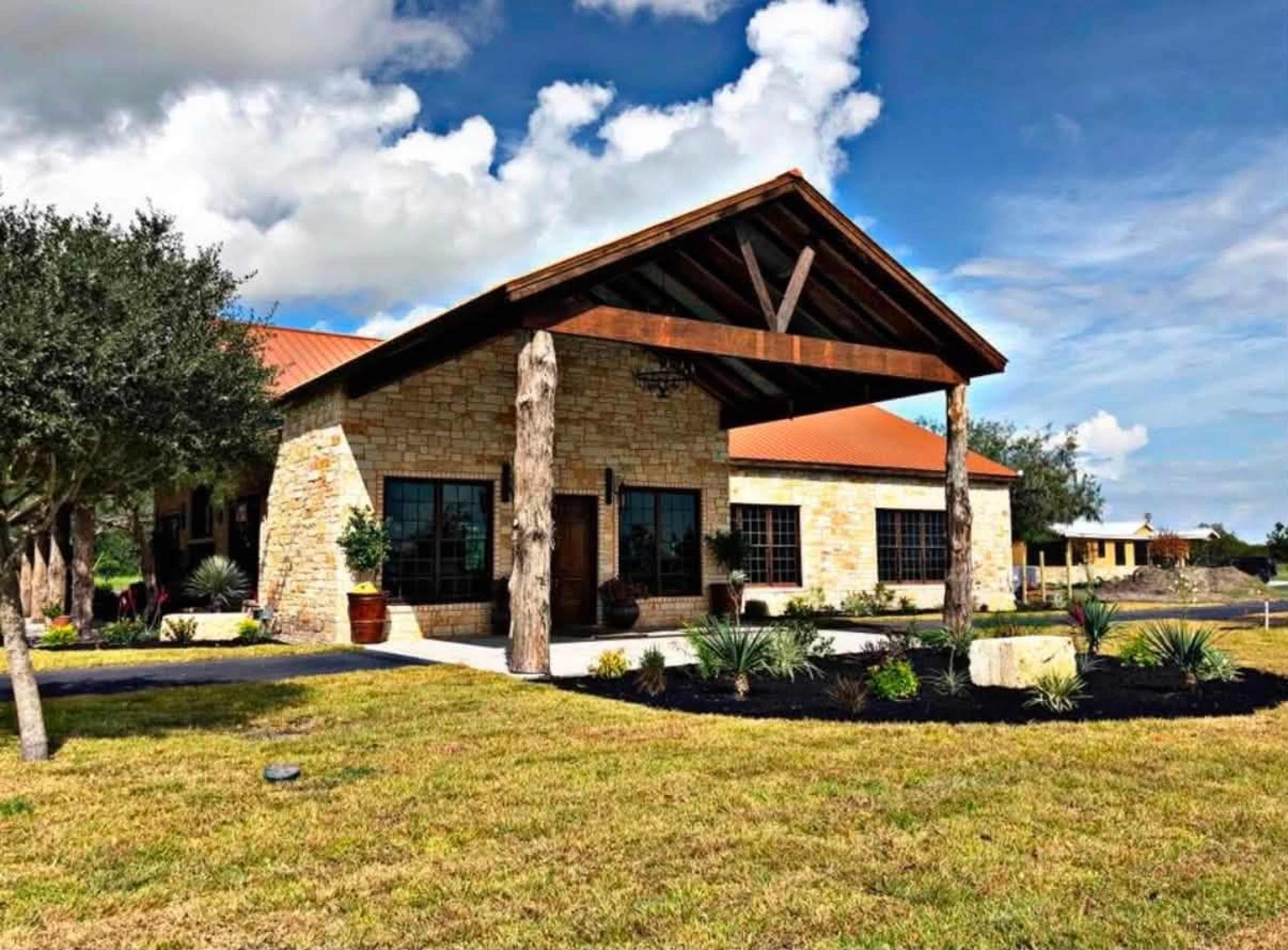 The image shows a large, modern stone house with a wooden porch and a red roof, surrounded by green grass and landscaping.