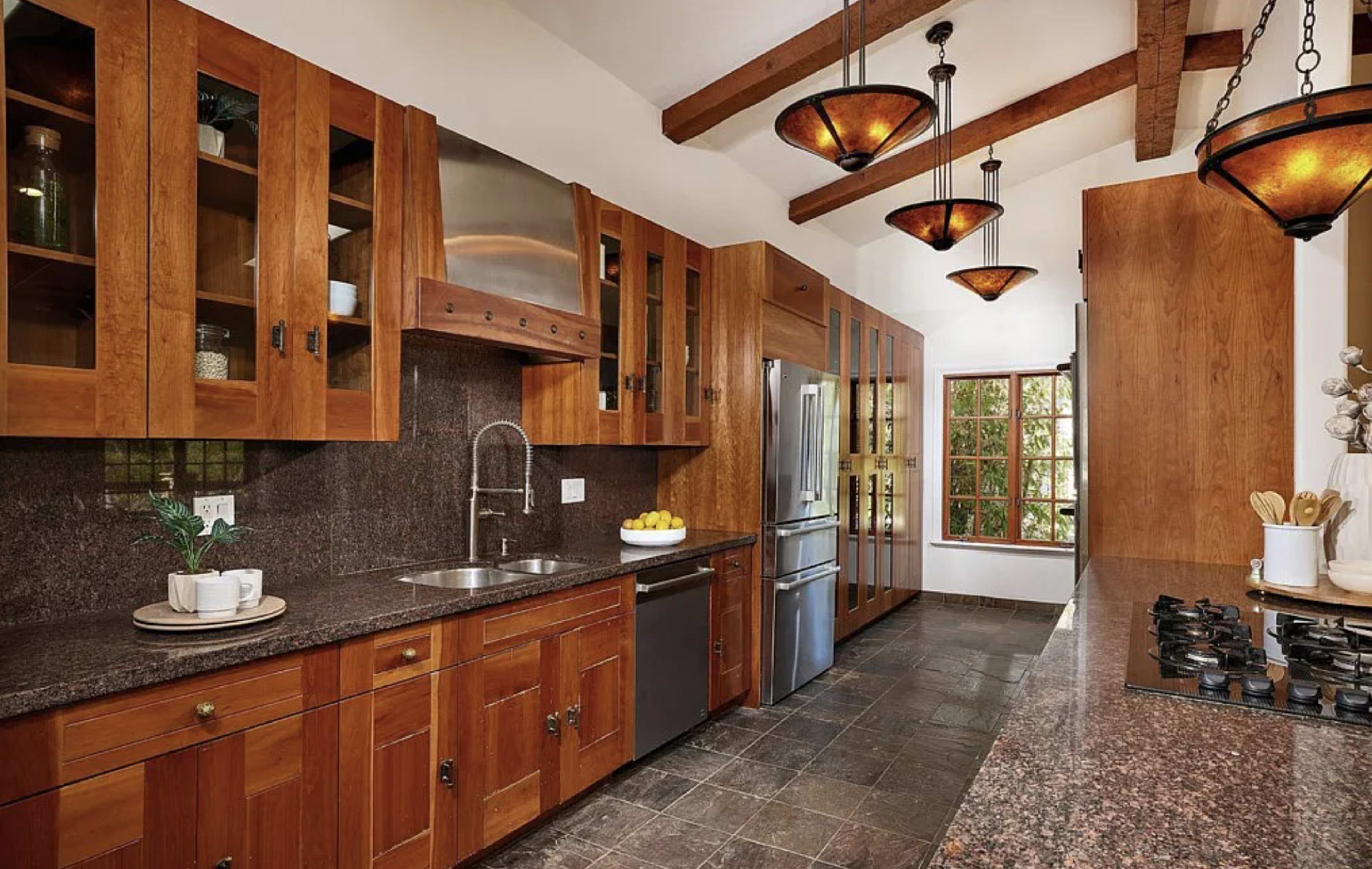 The image depicts a modern kitchen with wooden cabinetry, granite countertops, and stainless steel appliances, featuring pendant lighting and large windows.