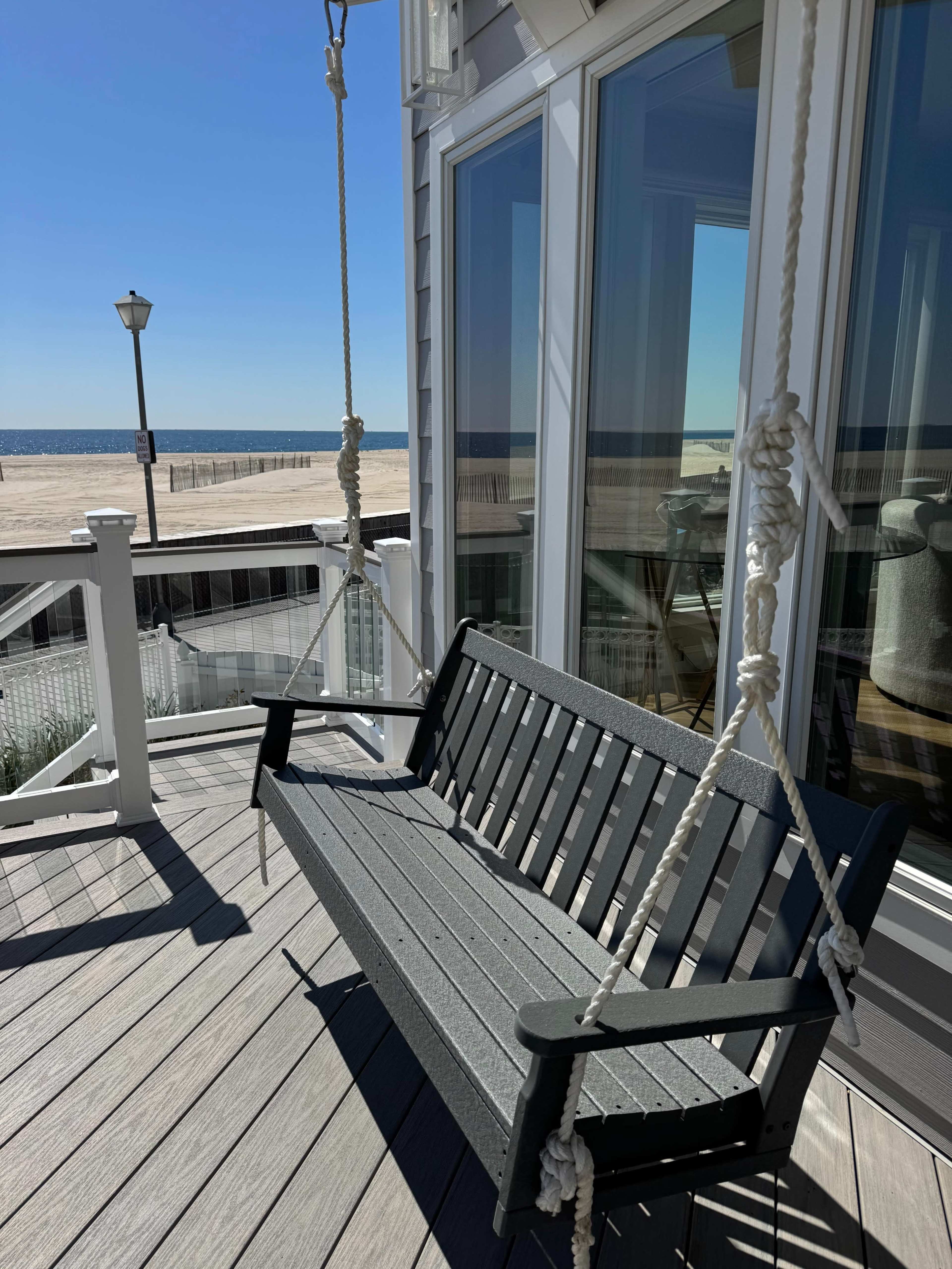 The image shows a swing bench hanging from ropes on a deck, with a view of a sandy beach and the ocean in the background.