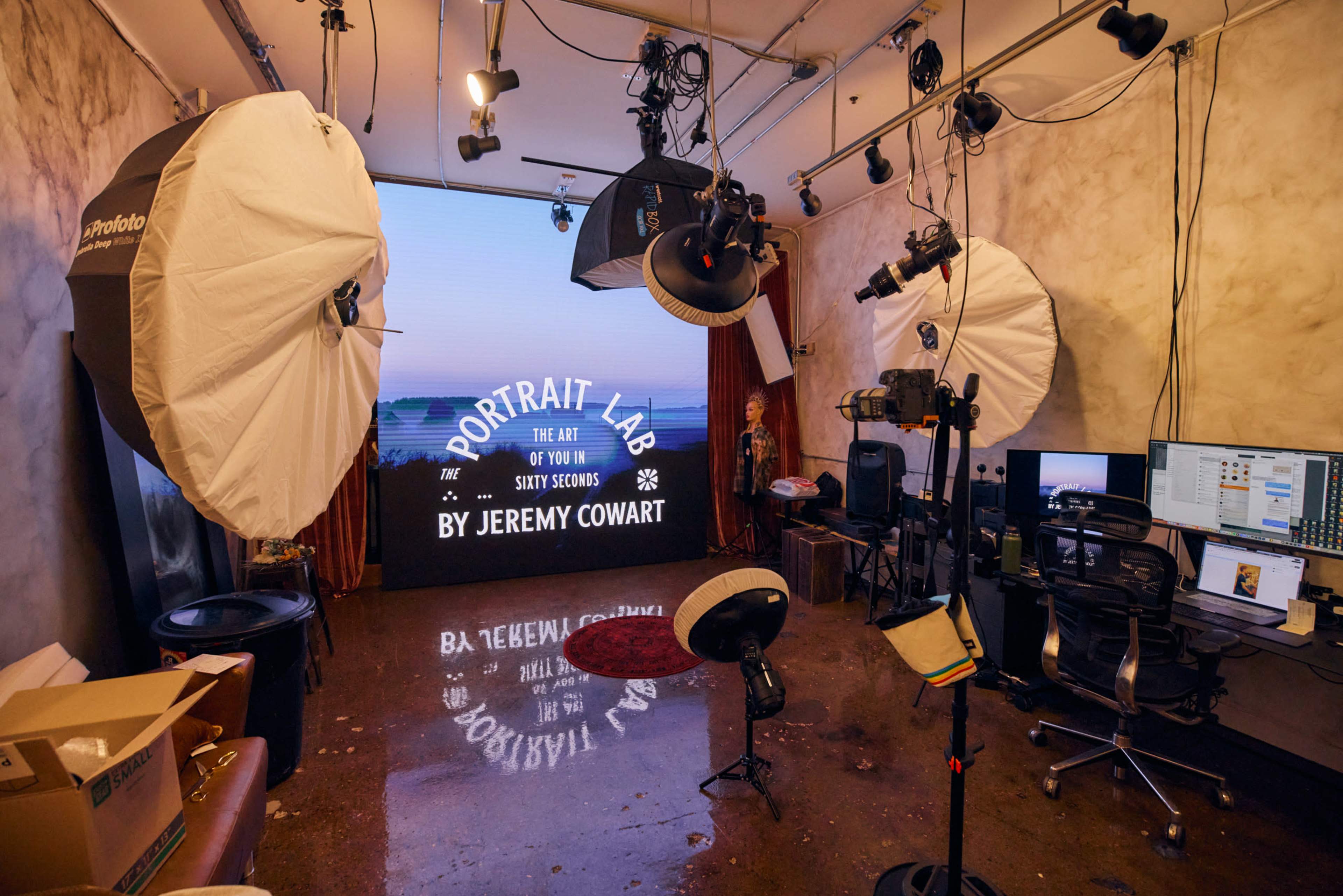 The image shows a photography studio equipped with lighting equipment, a large backdrop displaying "Portrait Lab" by Jeremy Cowart, and various camera setups alongside scattered boxes and furniture.