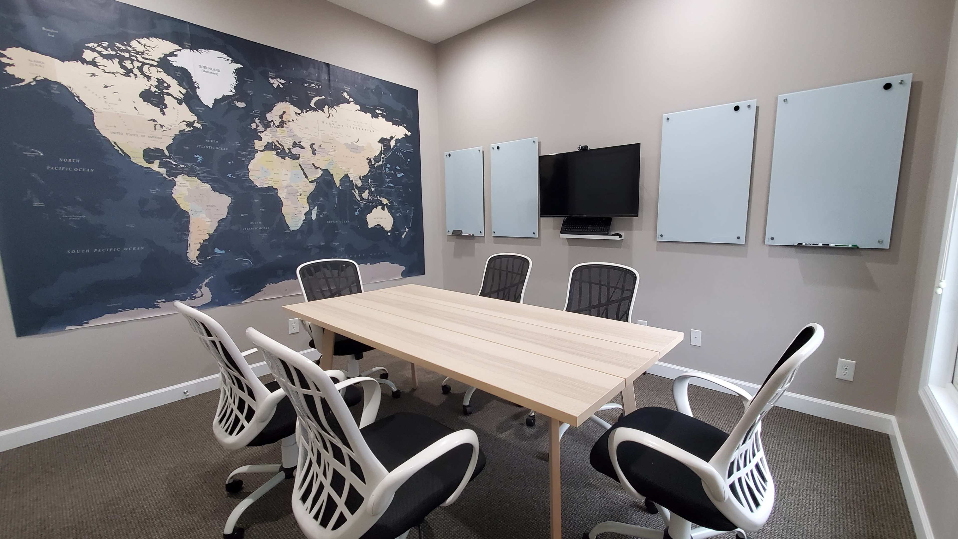 A conference room features a large wooden table surrounded by five chairs, with a world map on the wall and several whiteboards on either side.