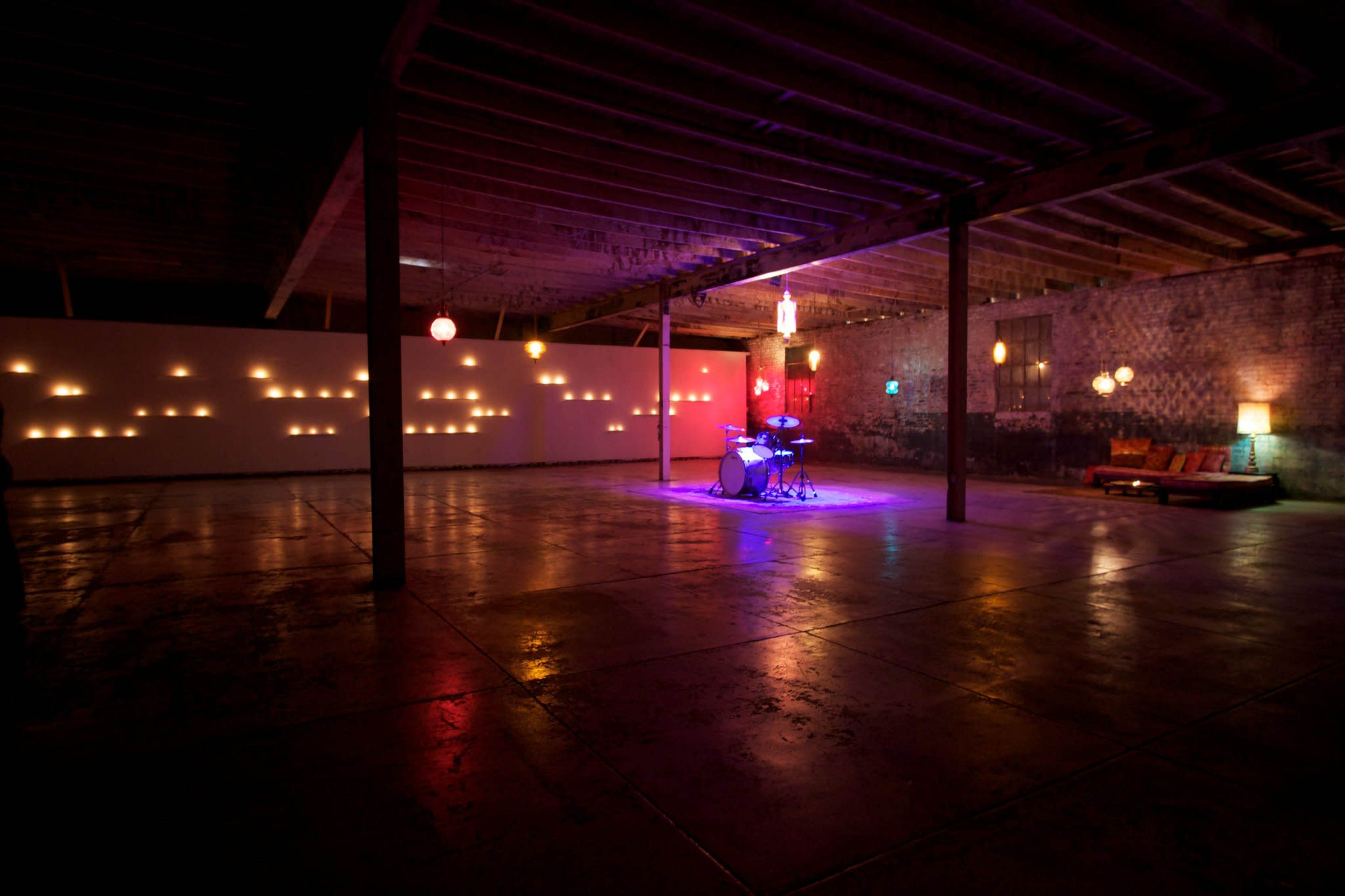 A spacious, dimly lit warehouse features a drum set illuminated by colored lights, with wall projections and hanging lamps casting soft glows around the room.
