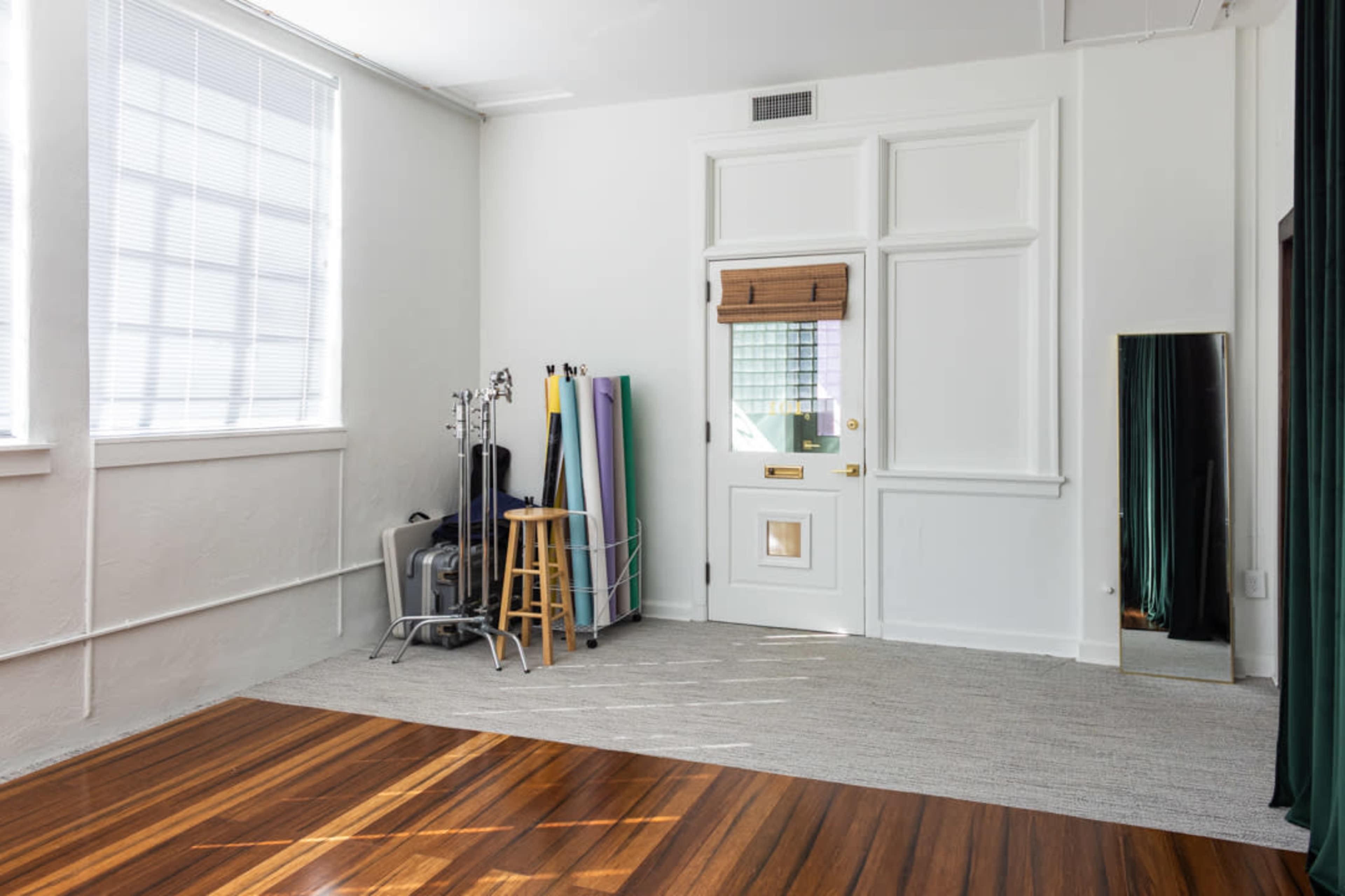 The image shows a bright, empty room with wooden flooring, a small door, a stool, and several colorful rolls of fabric against the wall.