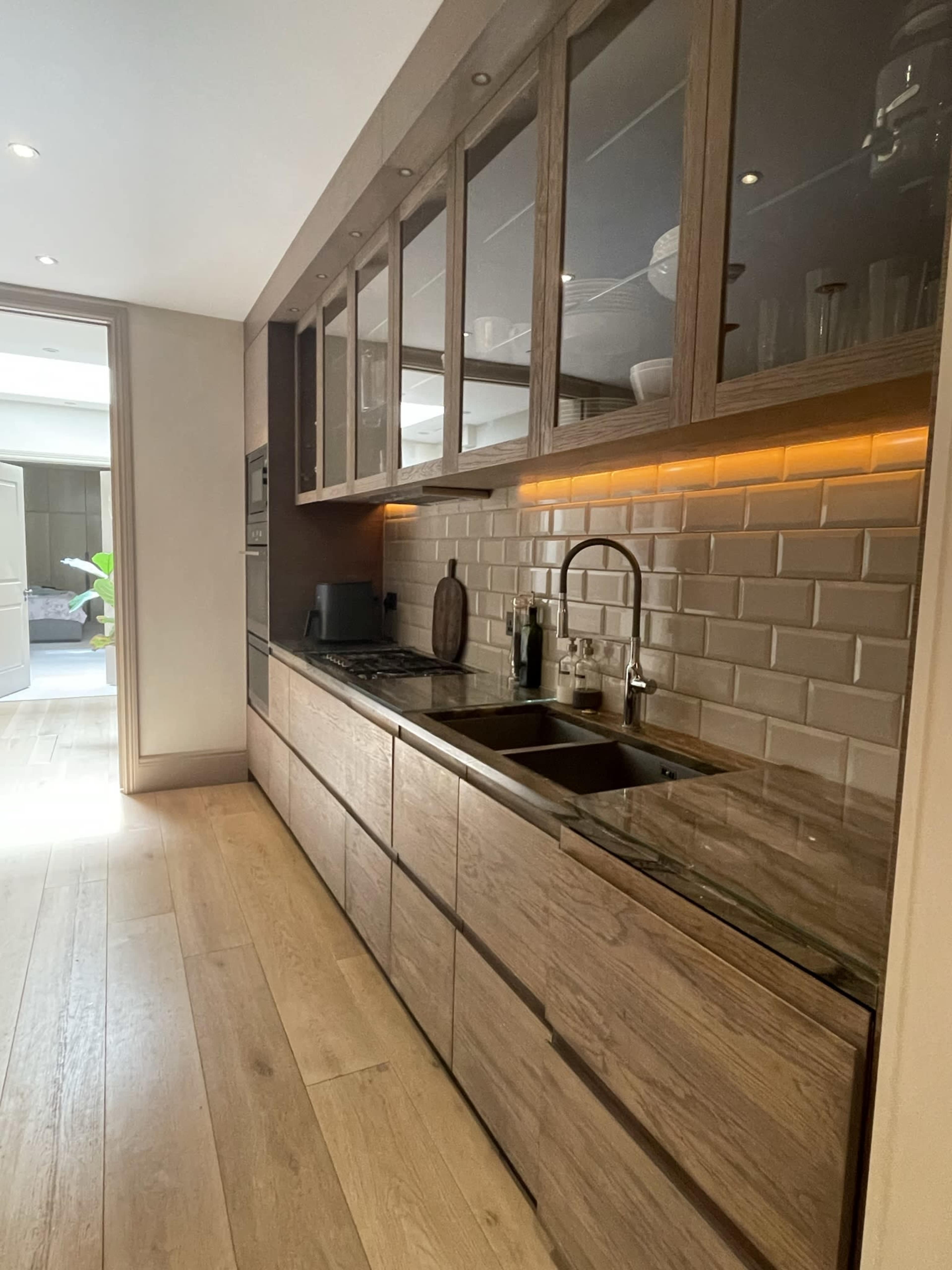A modern kitchen with wooden cabinetry, a sink, and a stone countertop, illuminated by under-cabinet lighting.