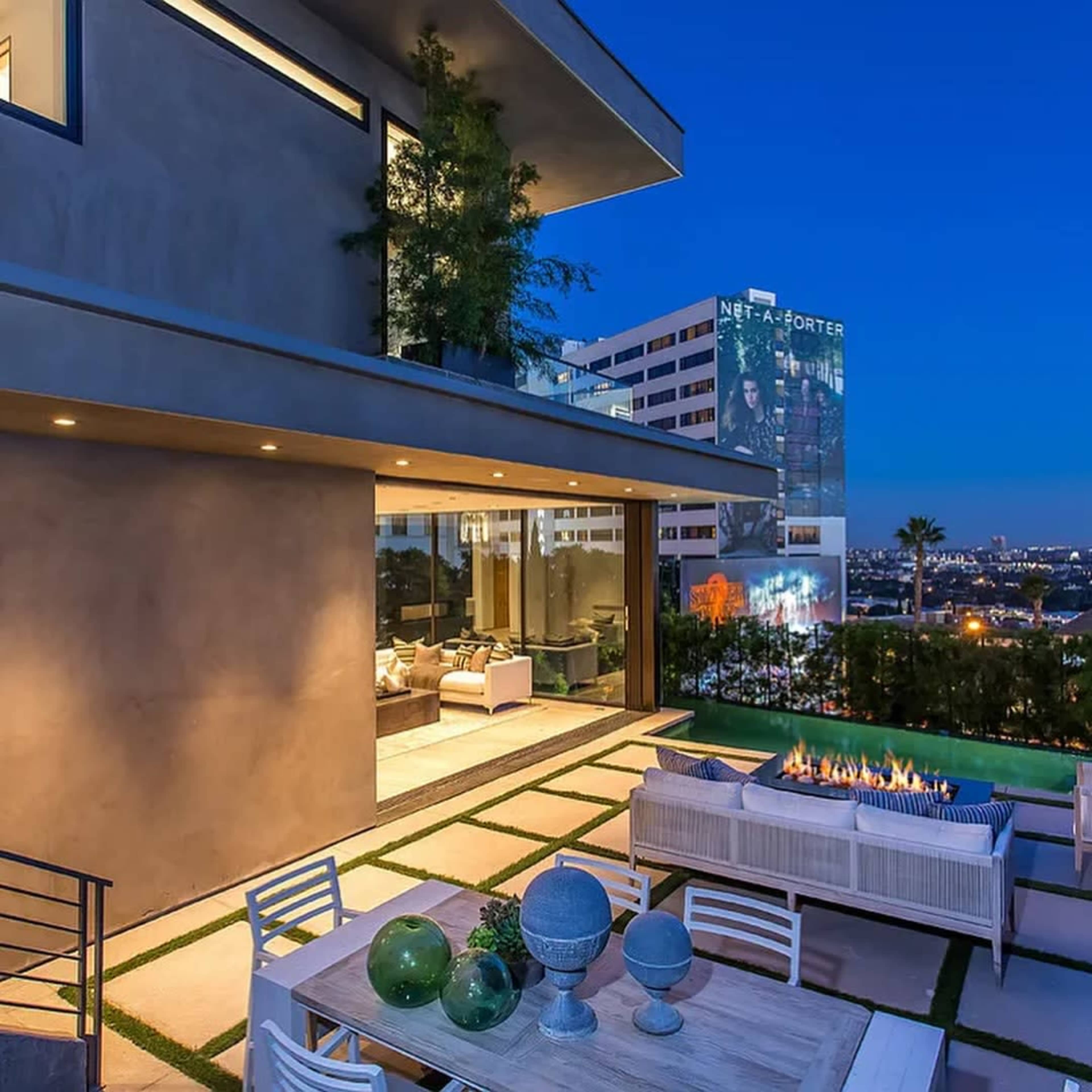 A modern outdoor living space features a table with decorative objects and a firepit, adjacent to a sleek building against a twilight sky.