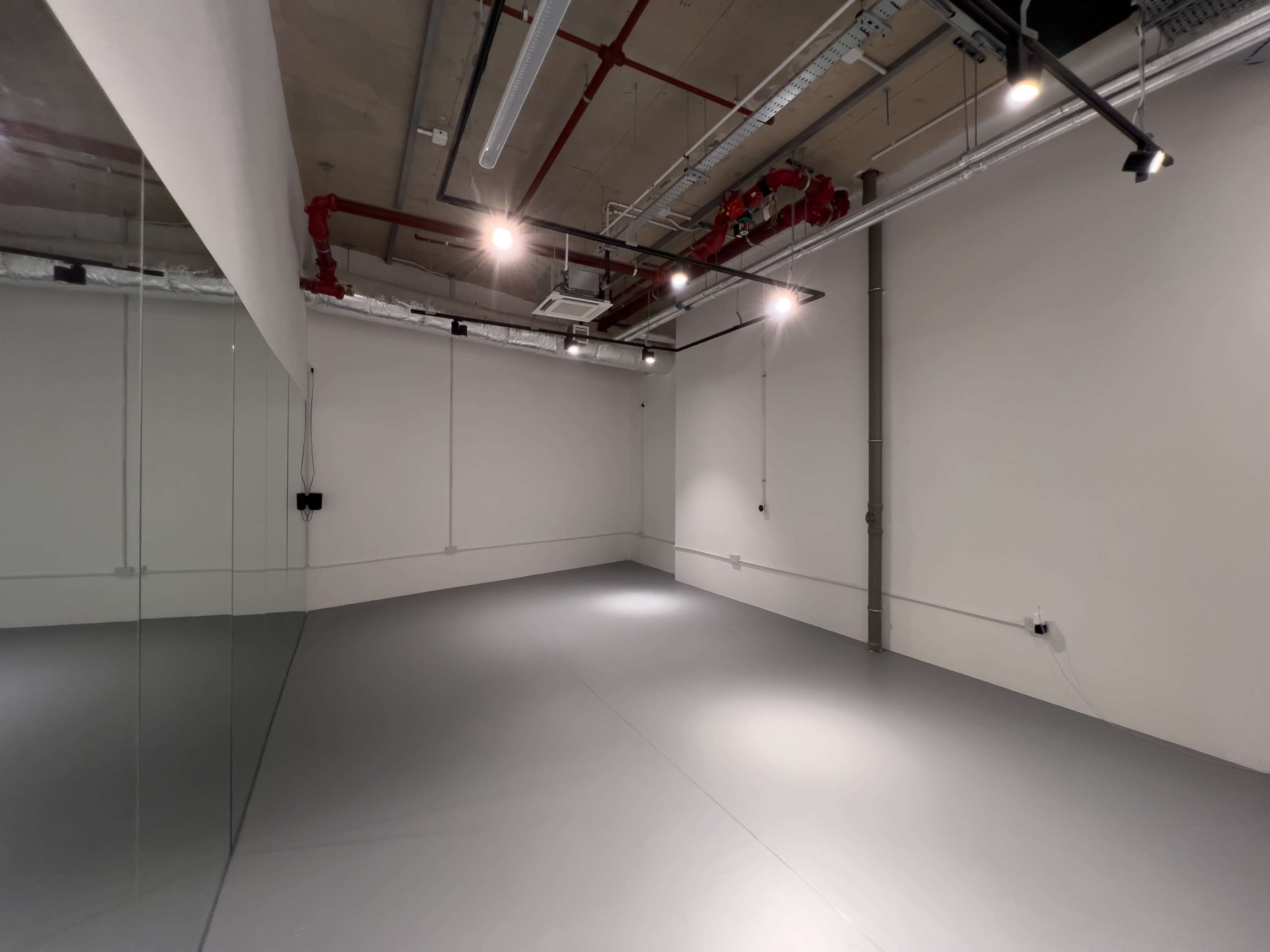 The image shows an empty room with gray flooring, concrete walls, and exposed ceiling pipes, illuminated by several overhead lights.