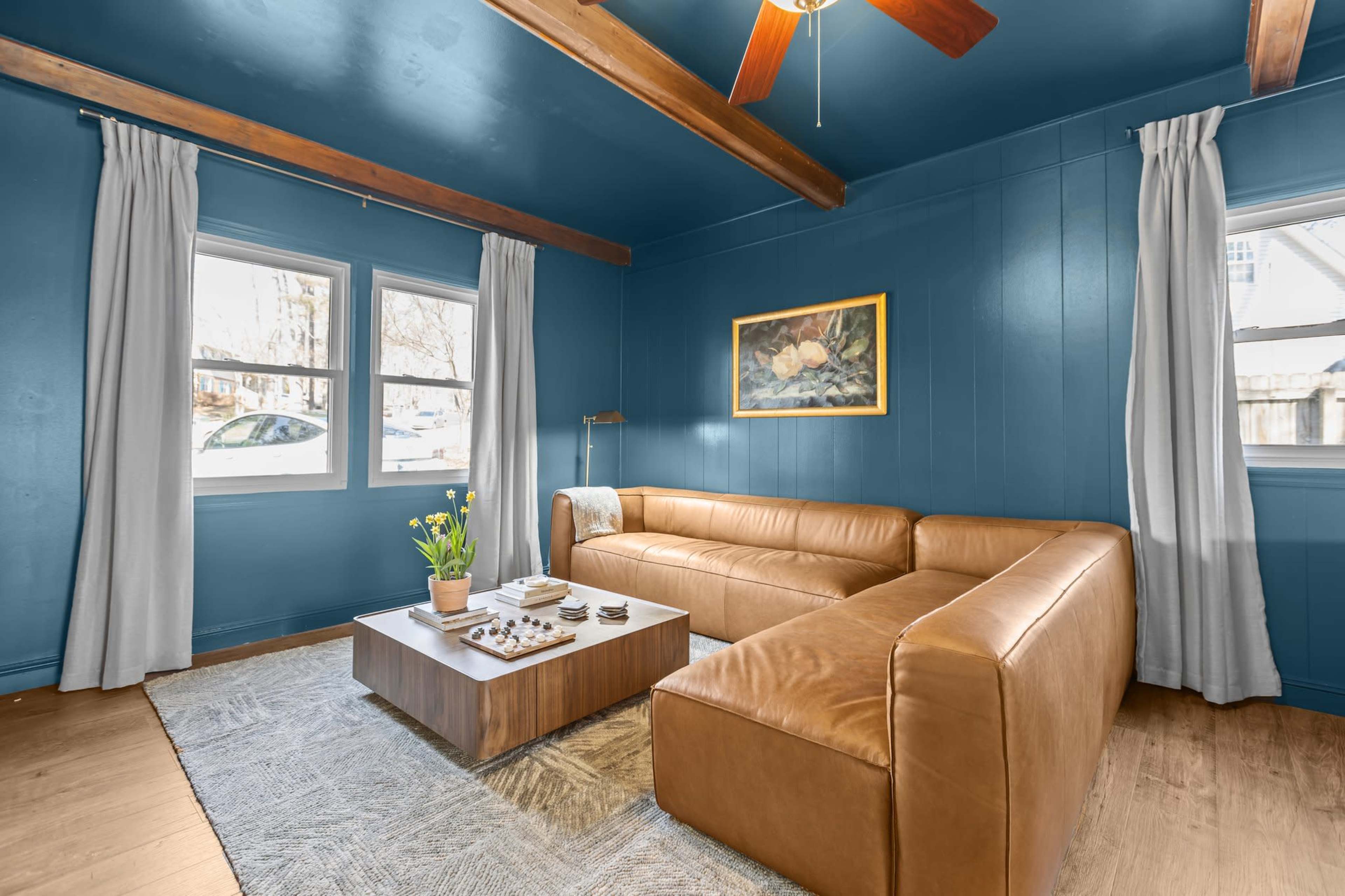 The living room features a brown leather sectional sofa, a wooden coffee table adorned with decorative items, and blue walls with natural light coming through two windows.