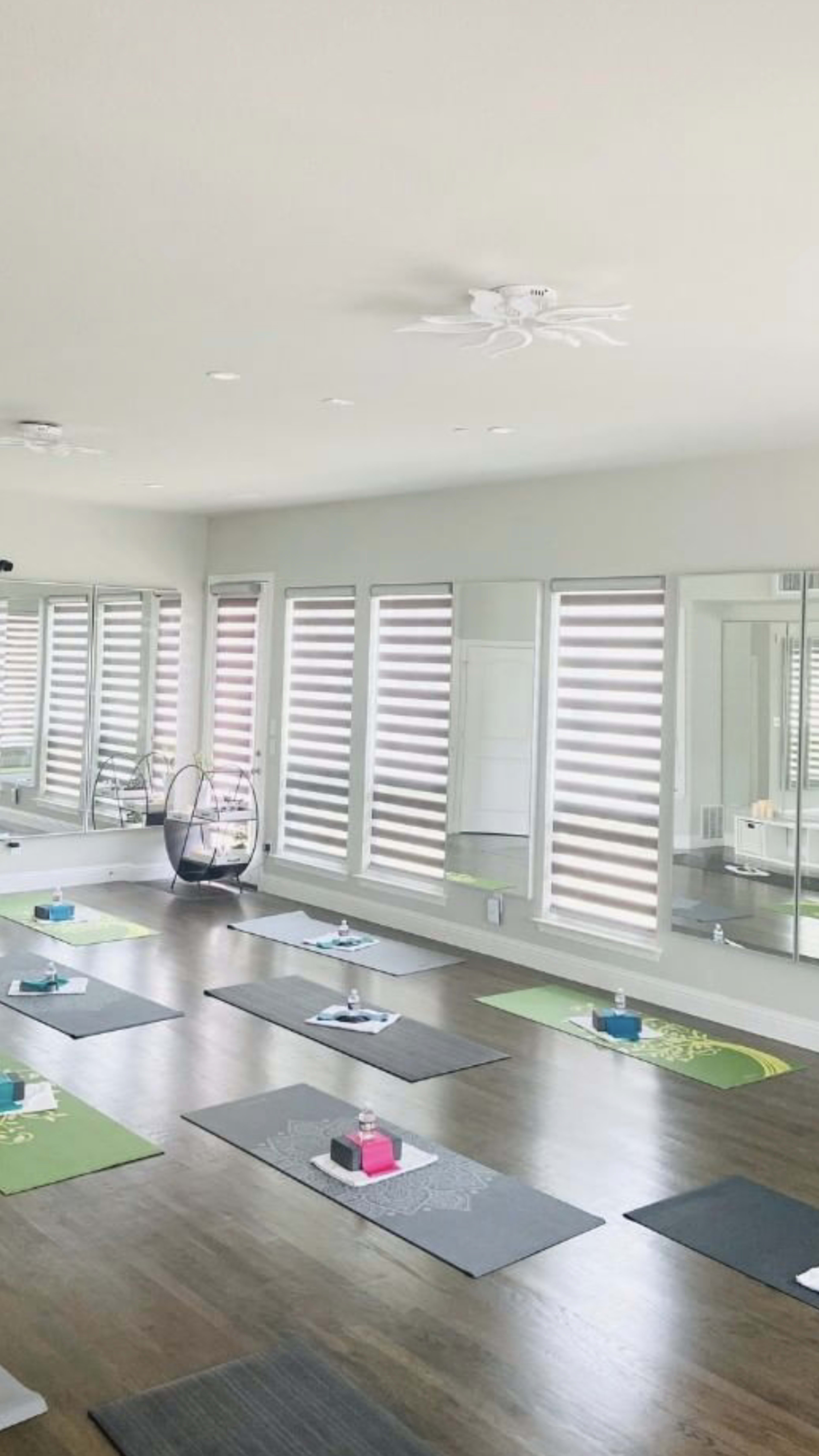 A spacious yoga studio with several yoga mats arranged on the wooden floor and large windows featuring horizontal blinds.