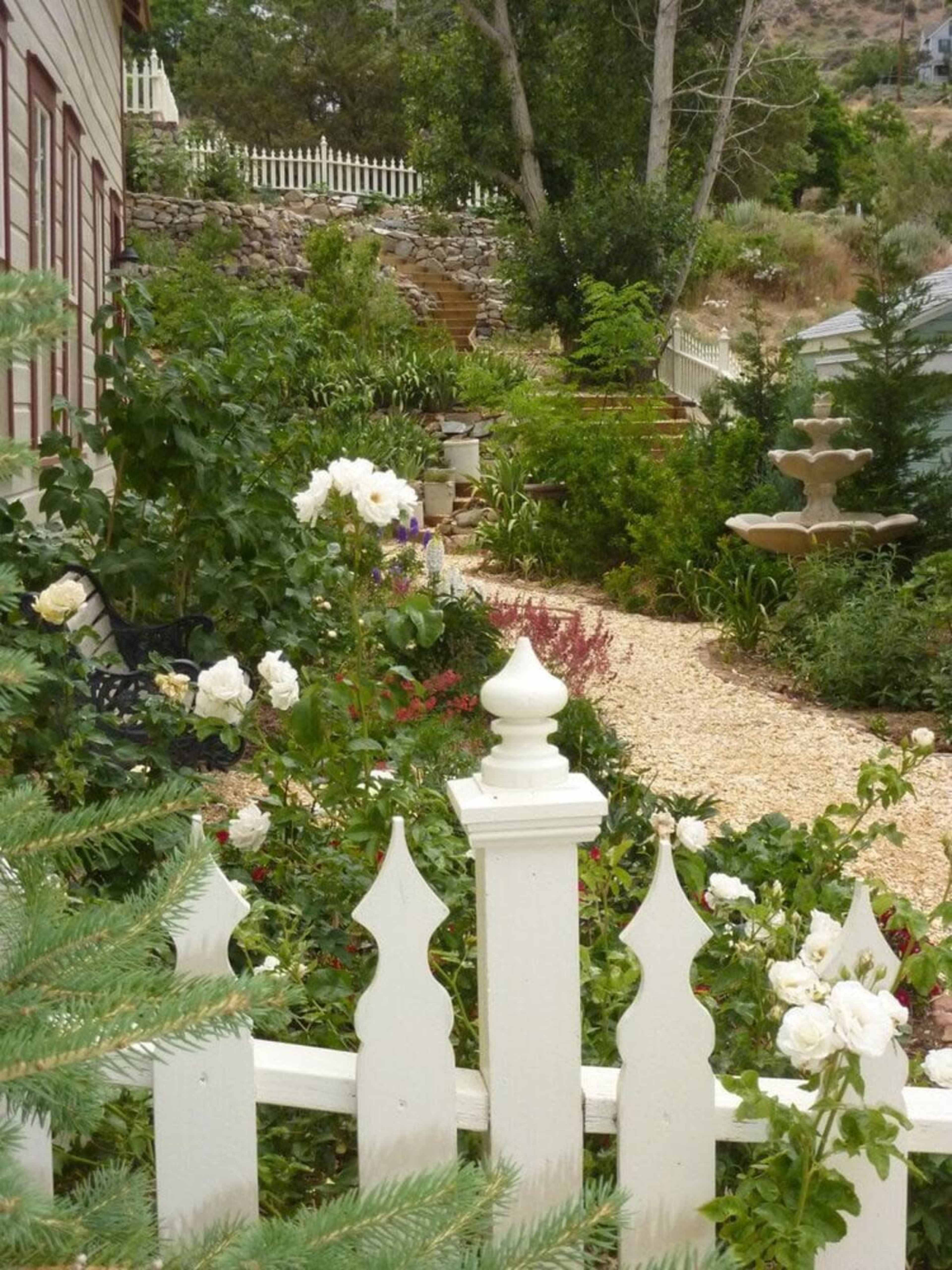 A white picket fence borders a gravel path that leads through a garden filled with various plants and flowers, featuring a fountain in the background.