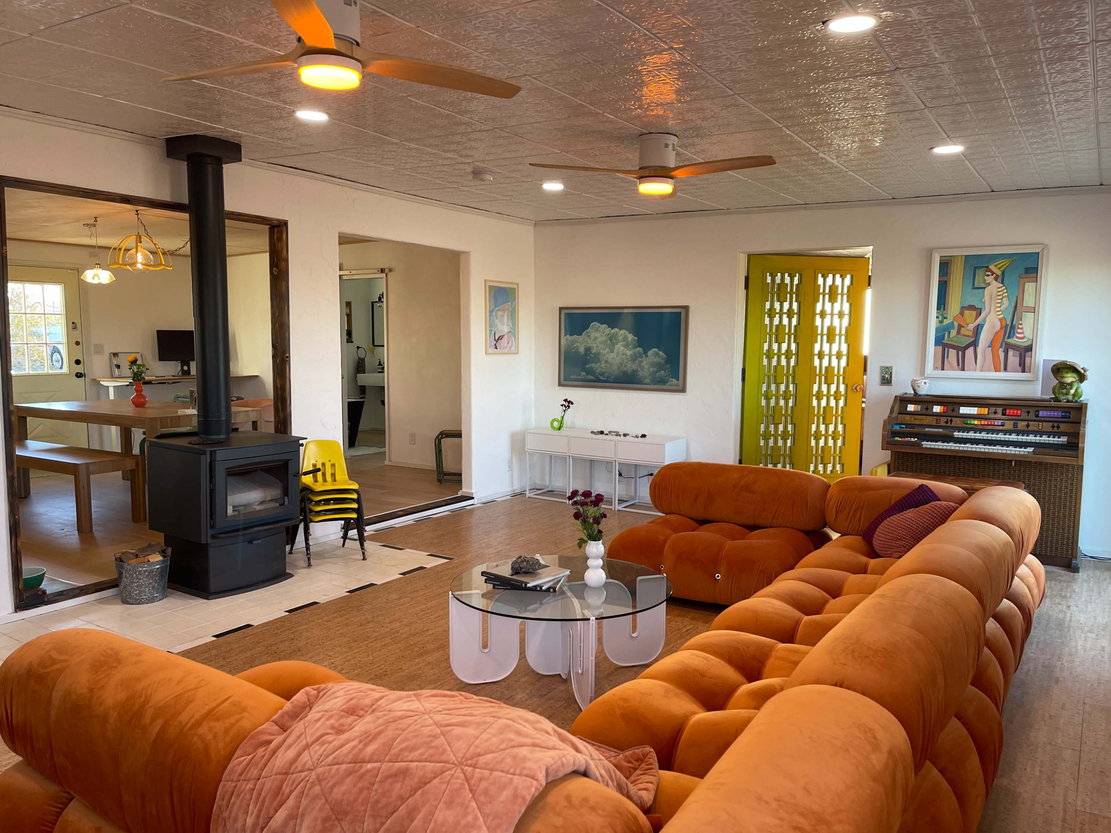 A bright living room features a large orange couch, a glass coffee table, a wood stove, and colorful artwork on the walls.