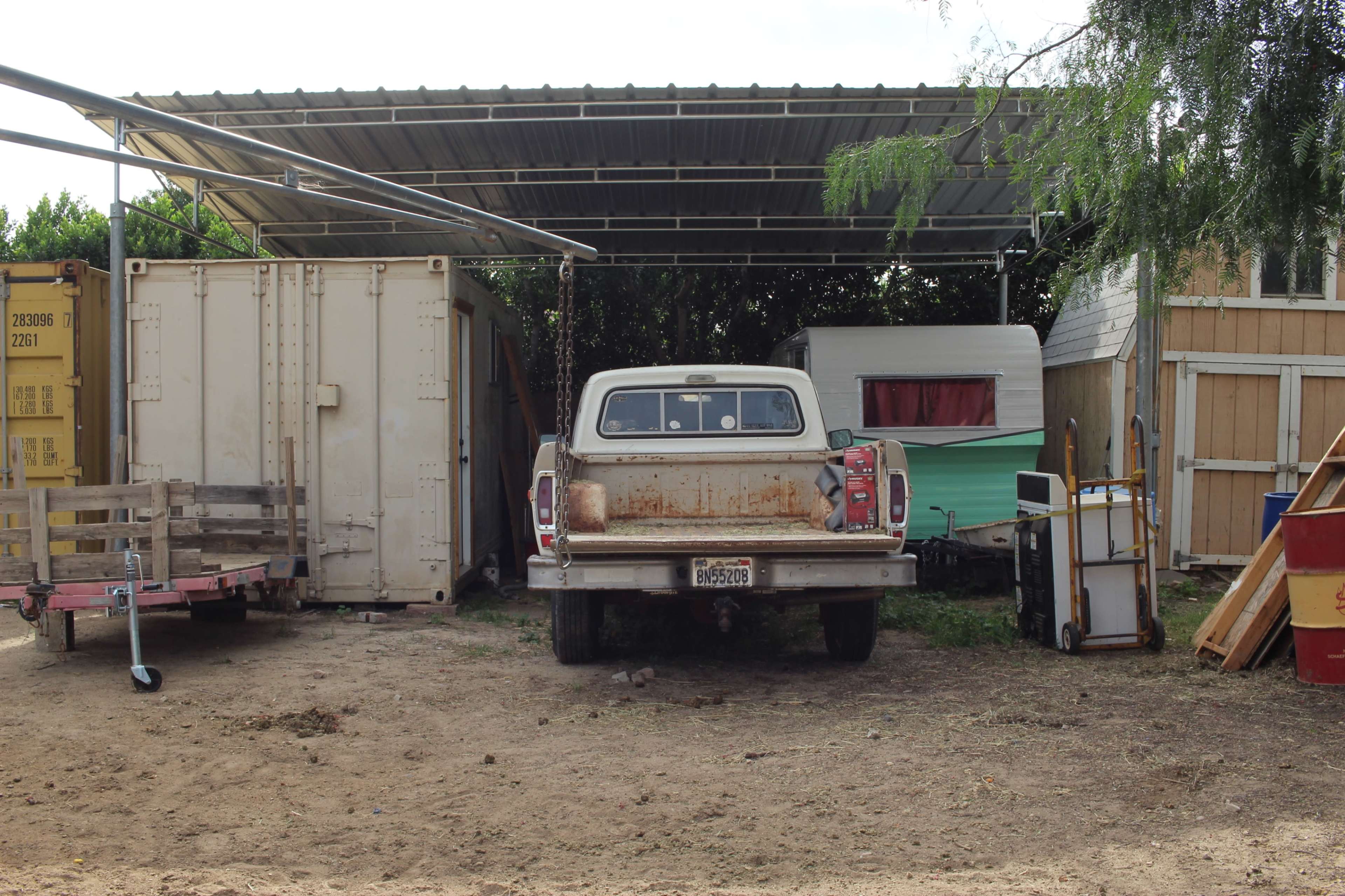 The image features a rusty pickup truck parked under a metal awning, surrounded by various storage containers and a small trailer.