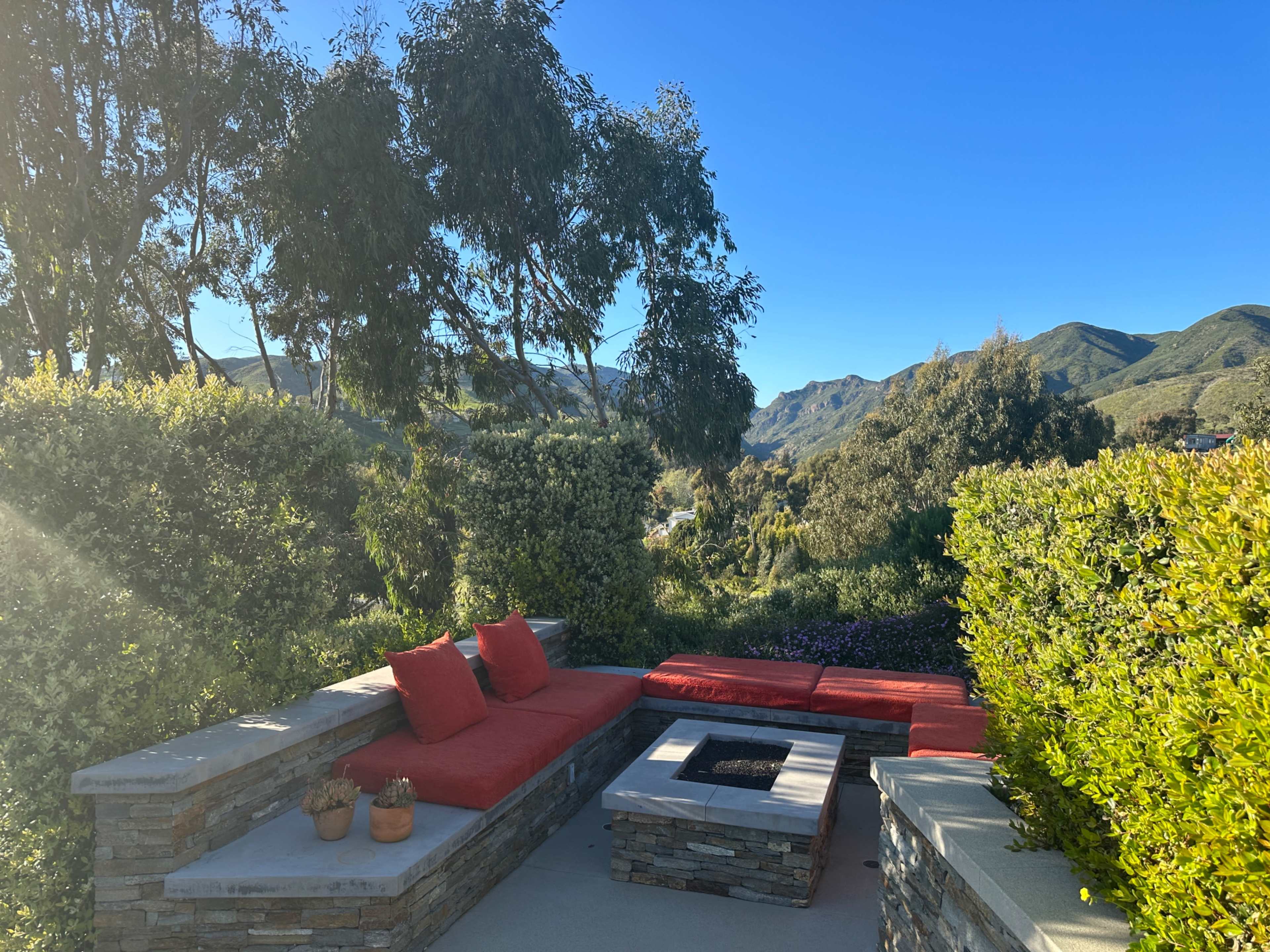 A stone seating area with red cushions surrounds a small fire pit, set against a backdrop of green hills and blue sky.