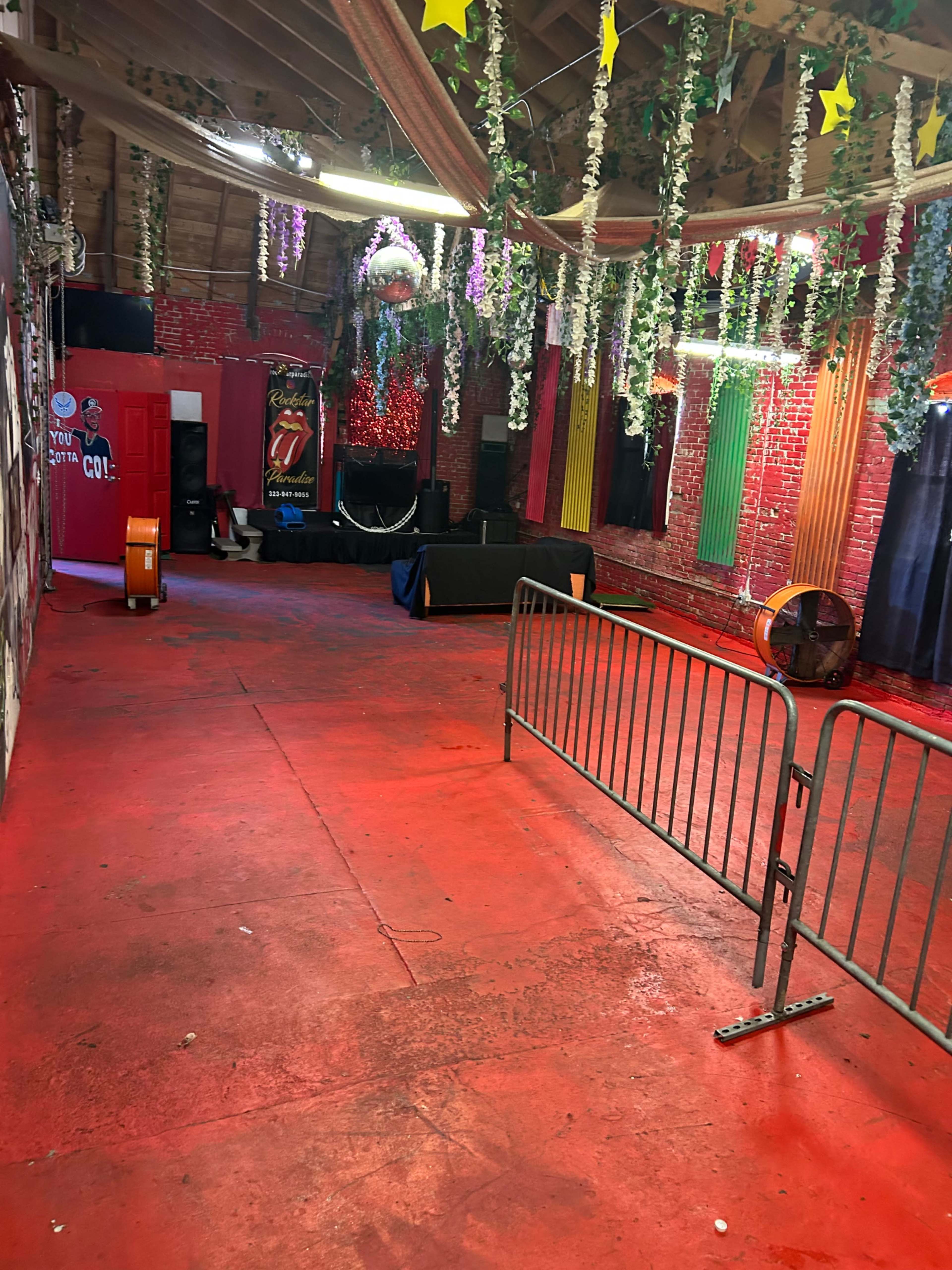 The image shows an empty, red-floored venue decorated with hanging plants and colorful curtains, featuring a stage area at the far end.