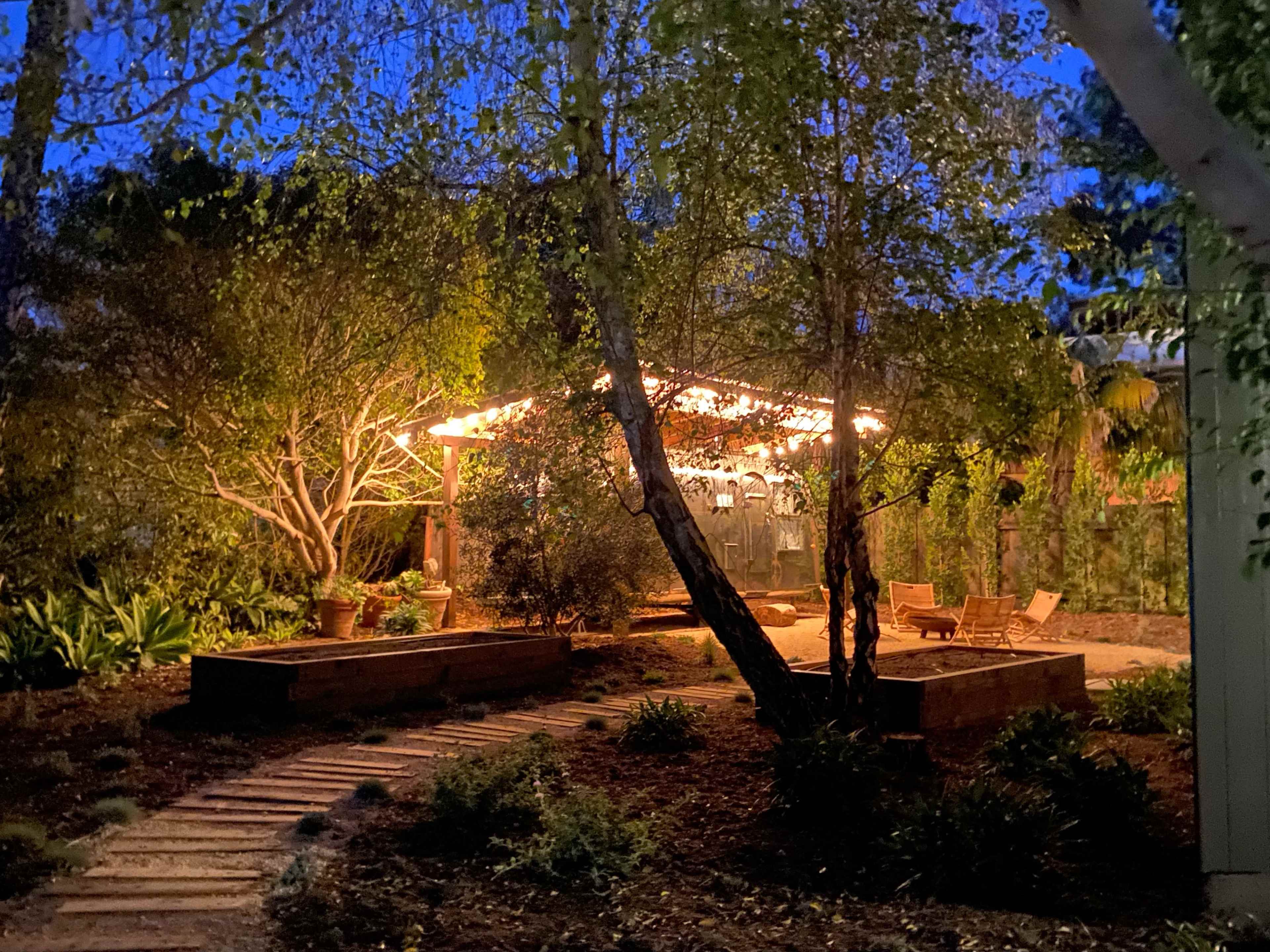 A pathway leads to a lit structure surrounded by trees and plants in a garden at night.