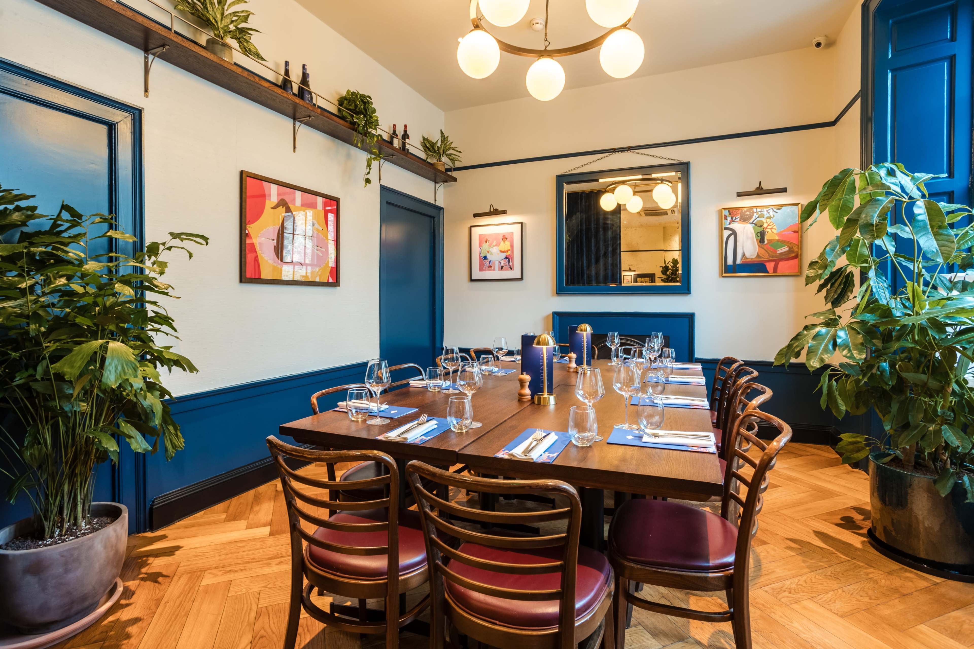 A dining area features a long wooden table set for a meal, surrounded by chairs and adorned with decorative plants and artwork on the walls.