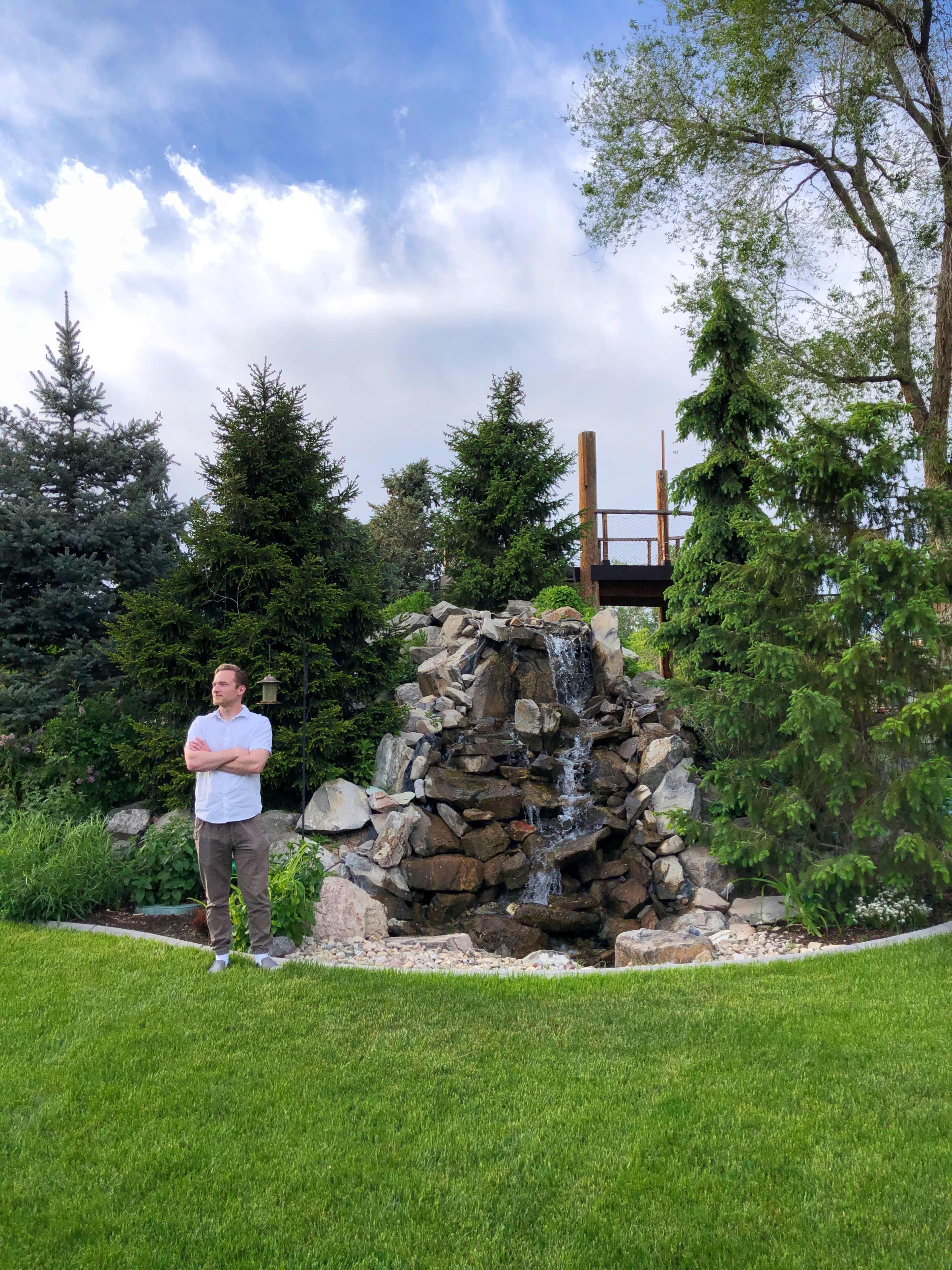 A man stands by a stone waterfall surrounded by greenery and trees, with a wooden deck in the background.