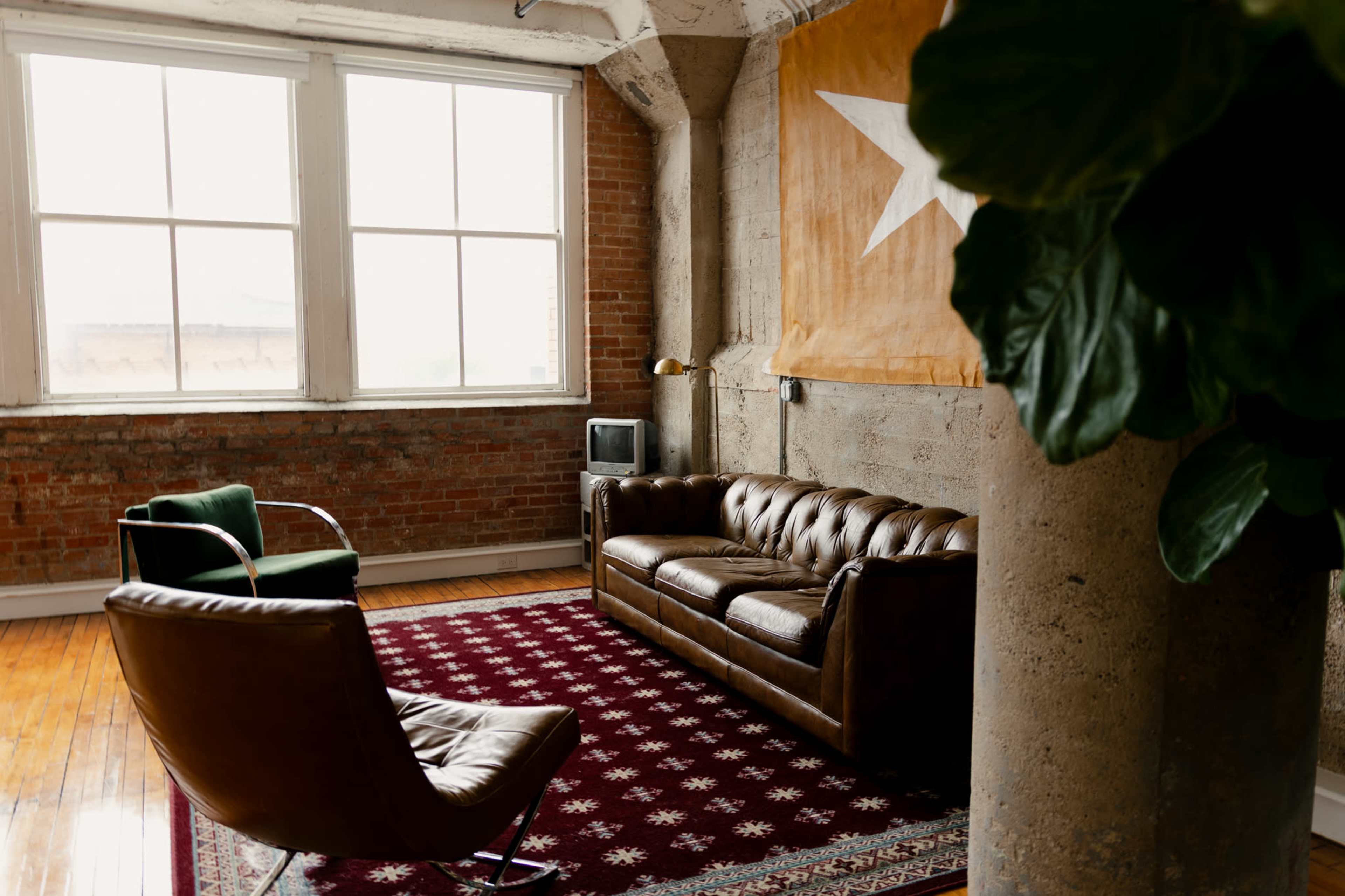 The image depicts a spacious, industrial-style living room featuring a brown leather sofa, a green armchair, and a vintage television near large windows with exposed brick walls and a star-patterned rug.