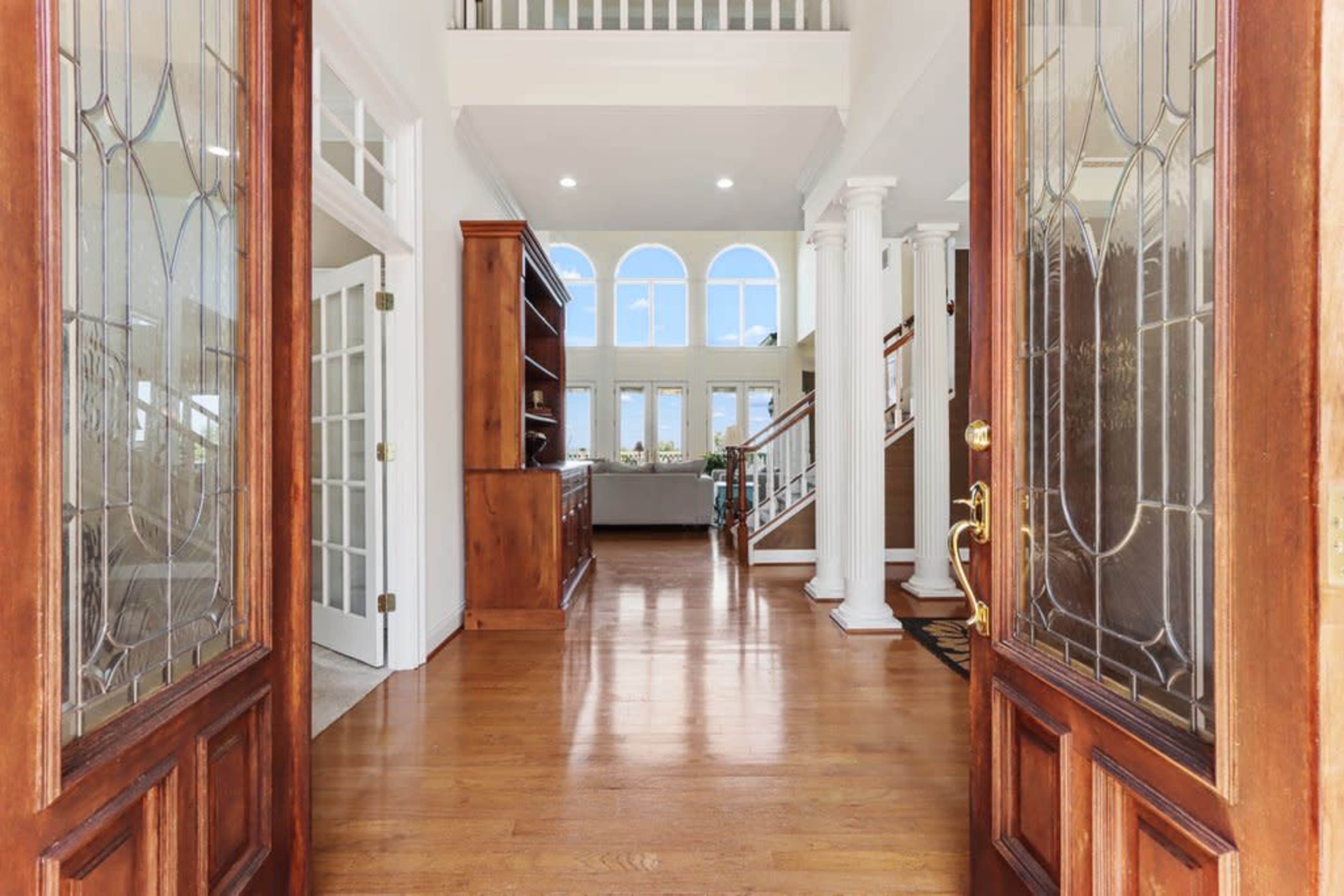 An entryway with double wooden doors opening to a spacious foyer featuring a second-level balcony and sunlight illuminating the hardwood floor.