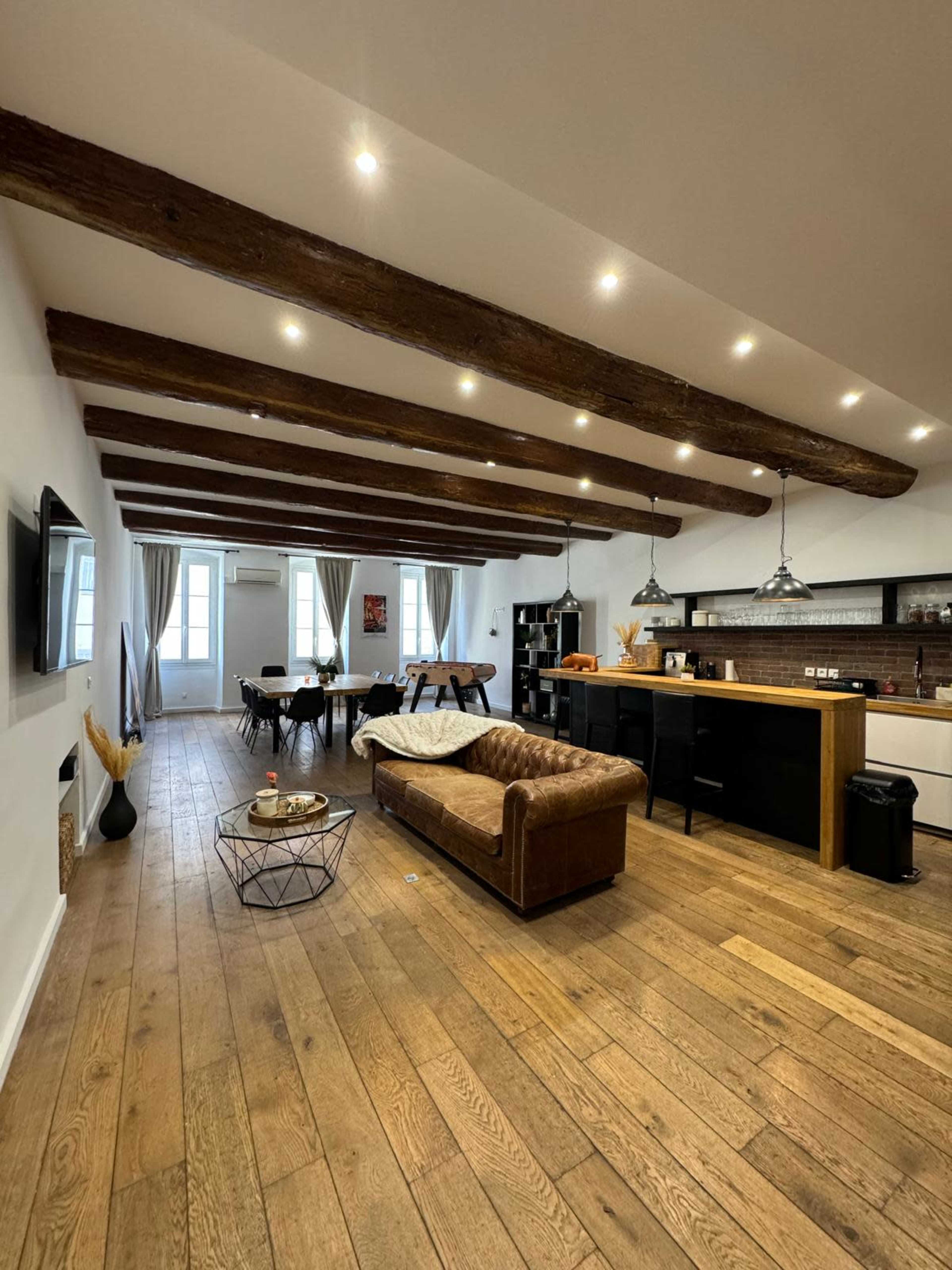 The image shows a modern open-concept living space with wooden beams, a brown leather sofa, and a dining area with a table and chairs.