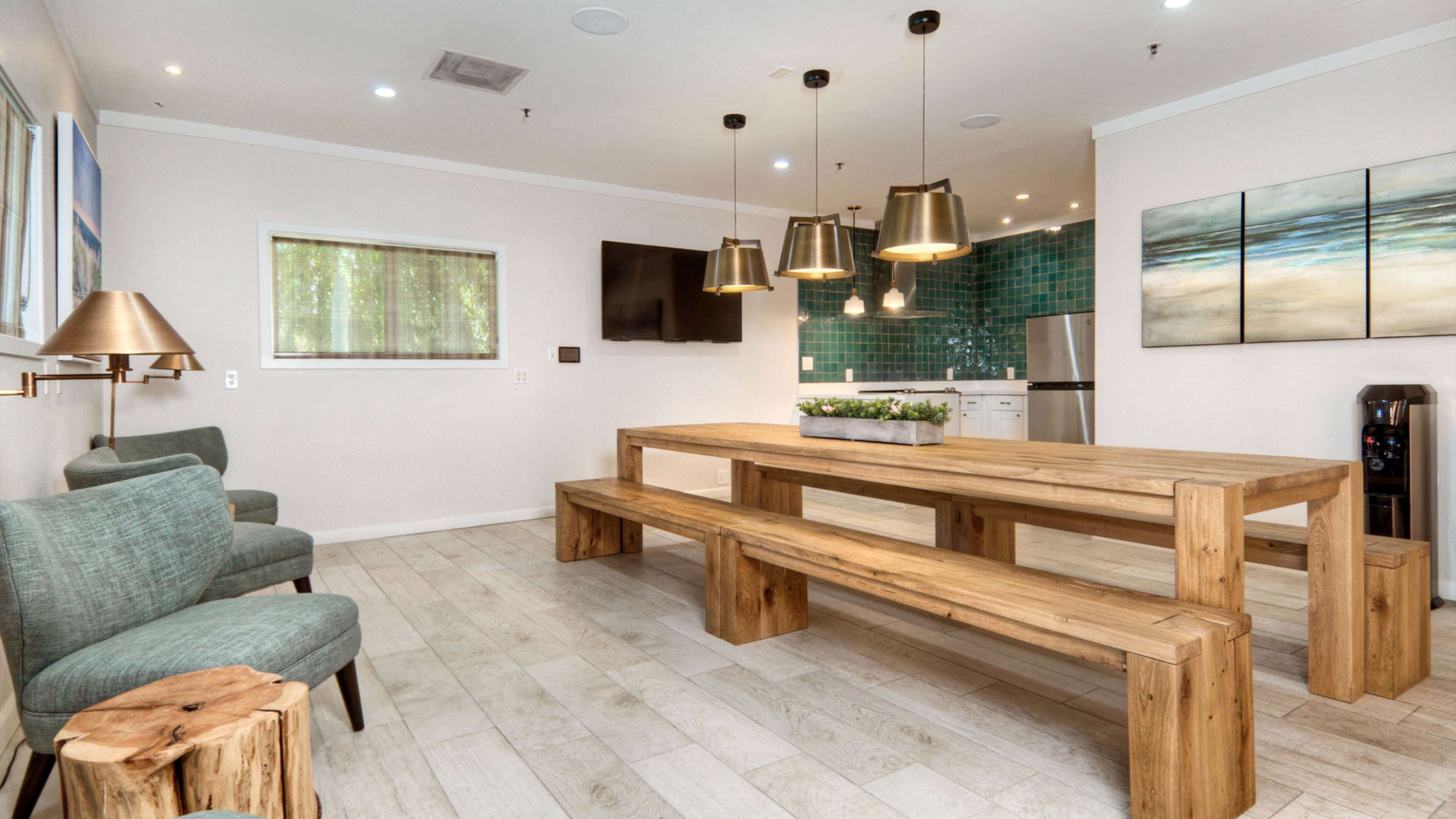 A modern dining area features a large wooden table with benches, surrounded by green upholstered chairs, and a kitchen area with teal tile in the background.