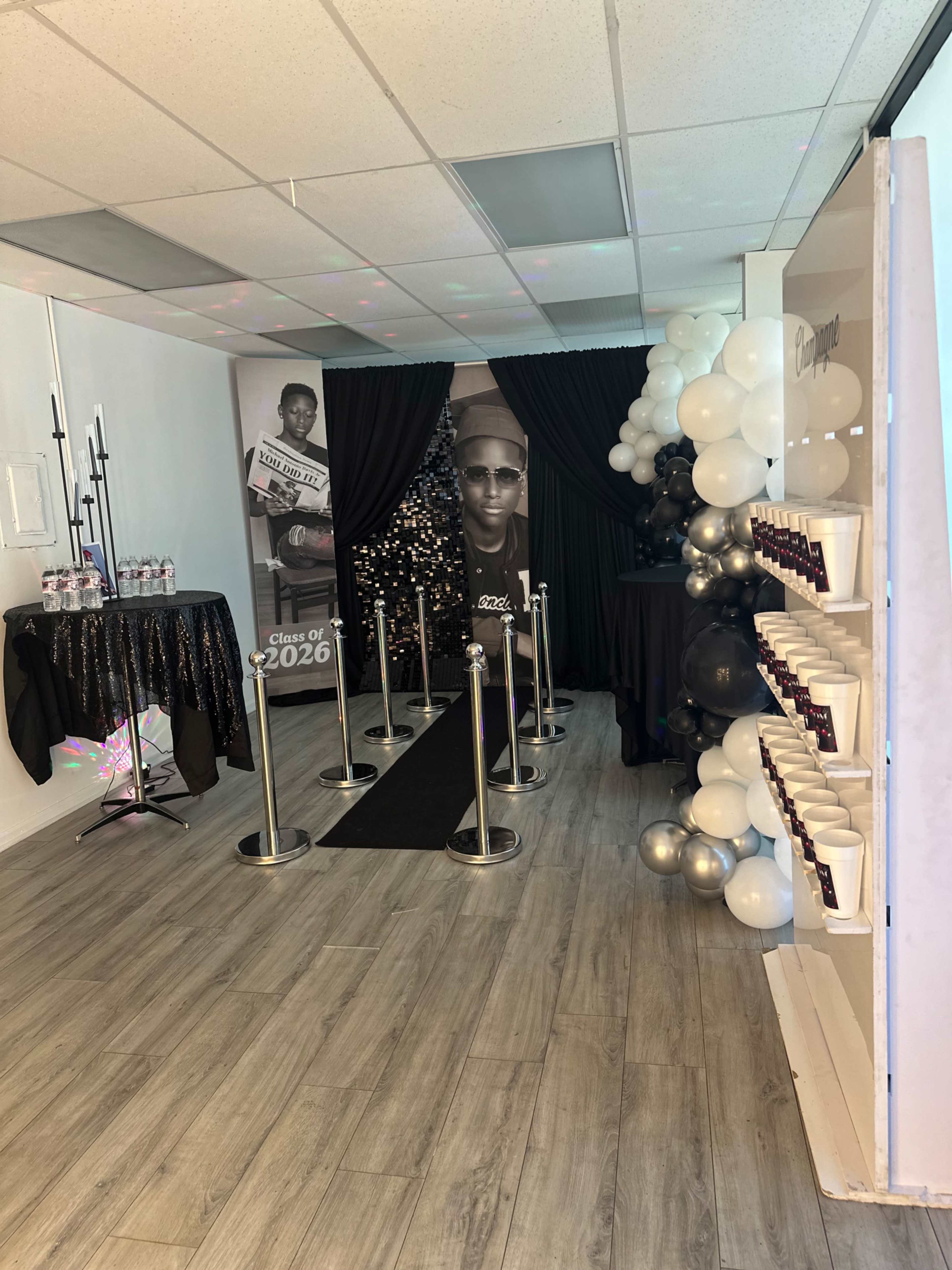 The image shows a decorated indoor space for a graduation celebration, featuring a black carpet runner, stanchions, a large poster of a graduate, and balloon arrangements in silver and black.