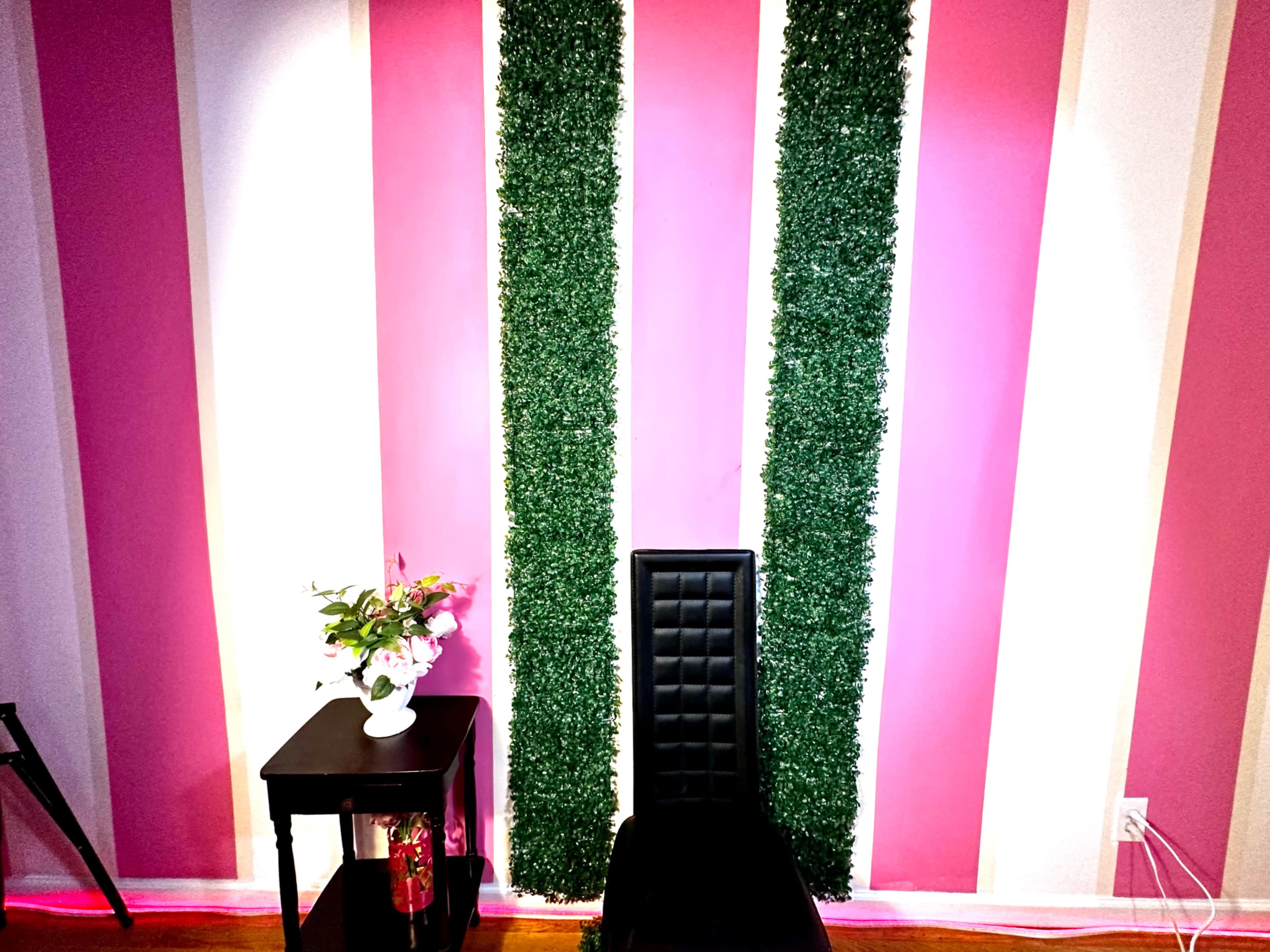 The image shows a corner of a room with pink striped walls, two vertical sections covered in green foliage, a black chair, and a small black table with a vase of flowers.