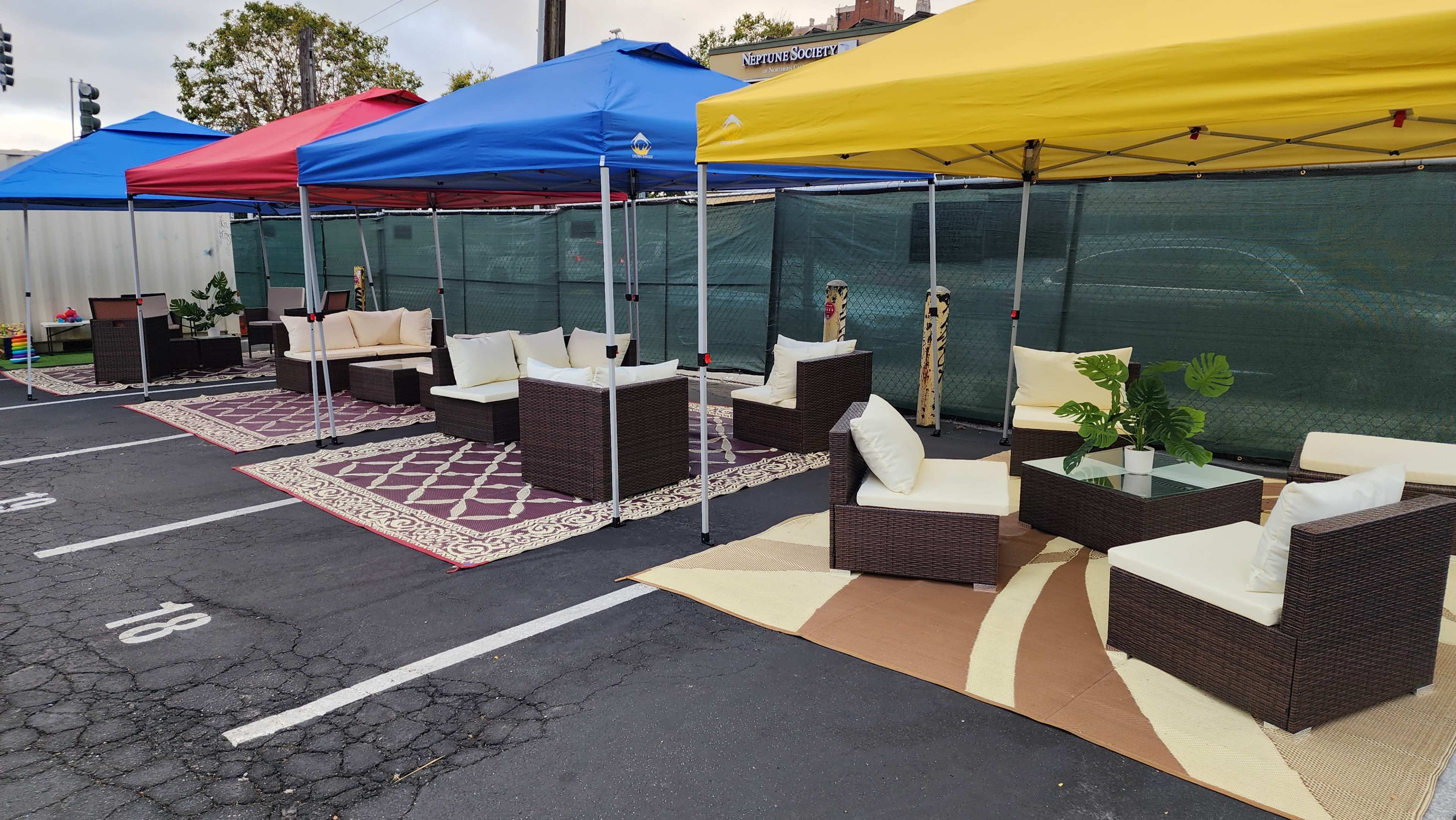 A series of outdoor seating arrangements under colorful canopies are set up on a parking lot, featuring couches and decorative rugs.