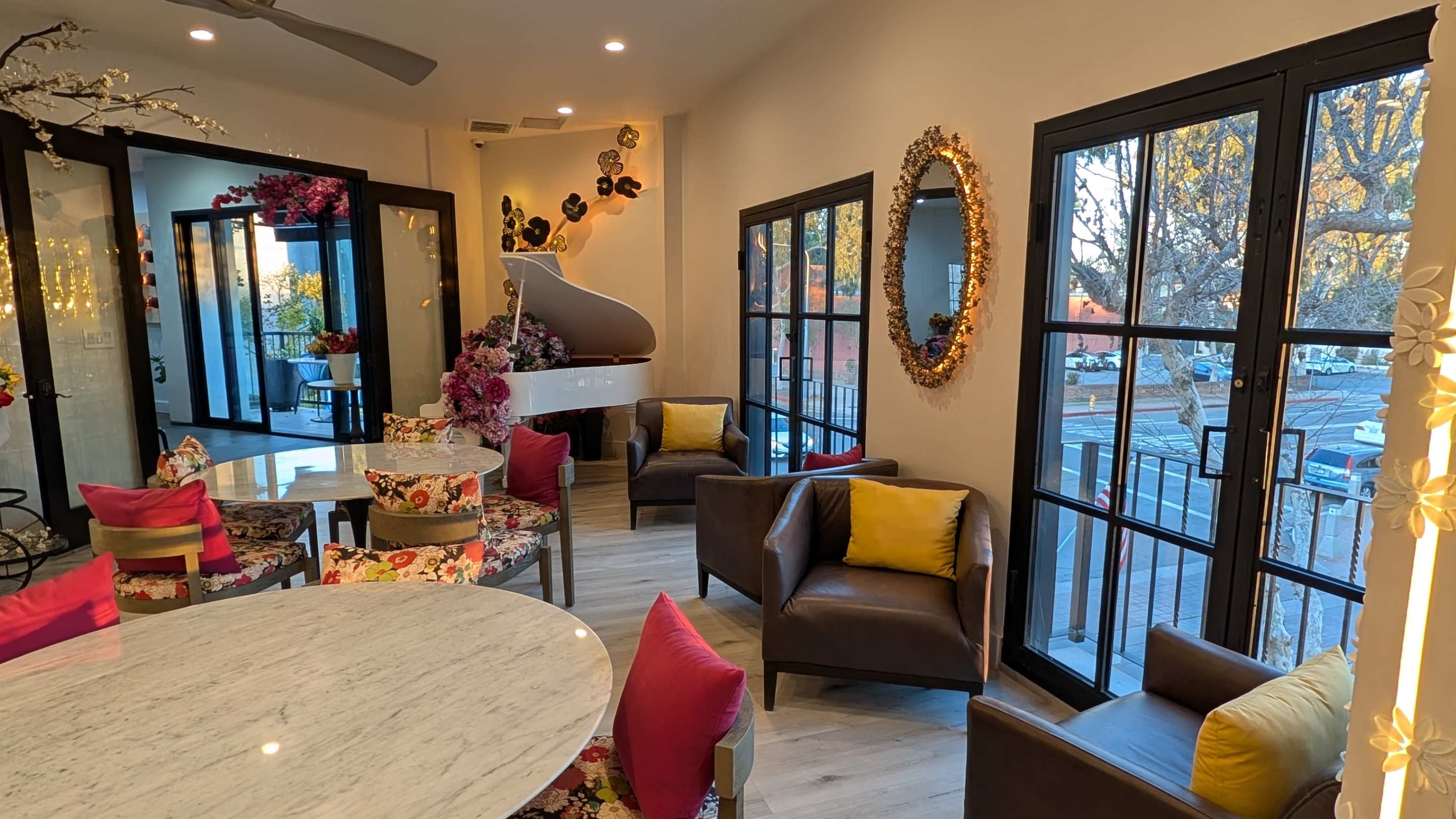 The image shows a well-lit café interior featuring a white piano, colorful floral-patterned chairs, and large windows overlooking an outdoor view.