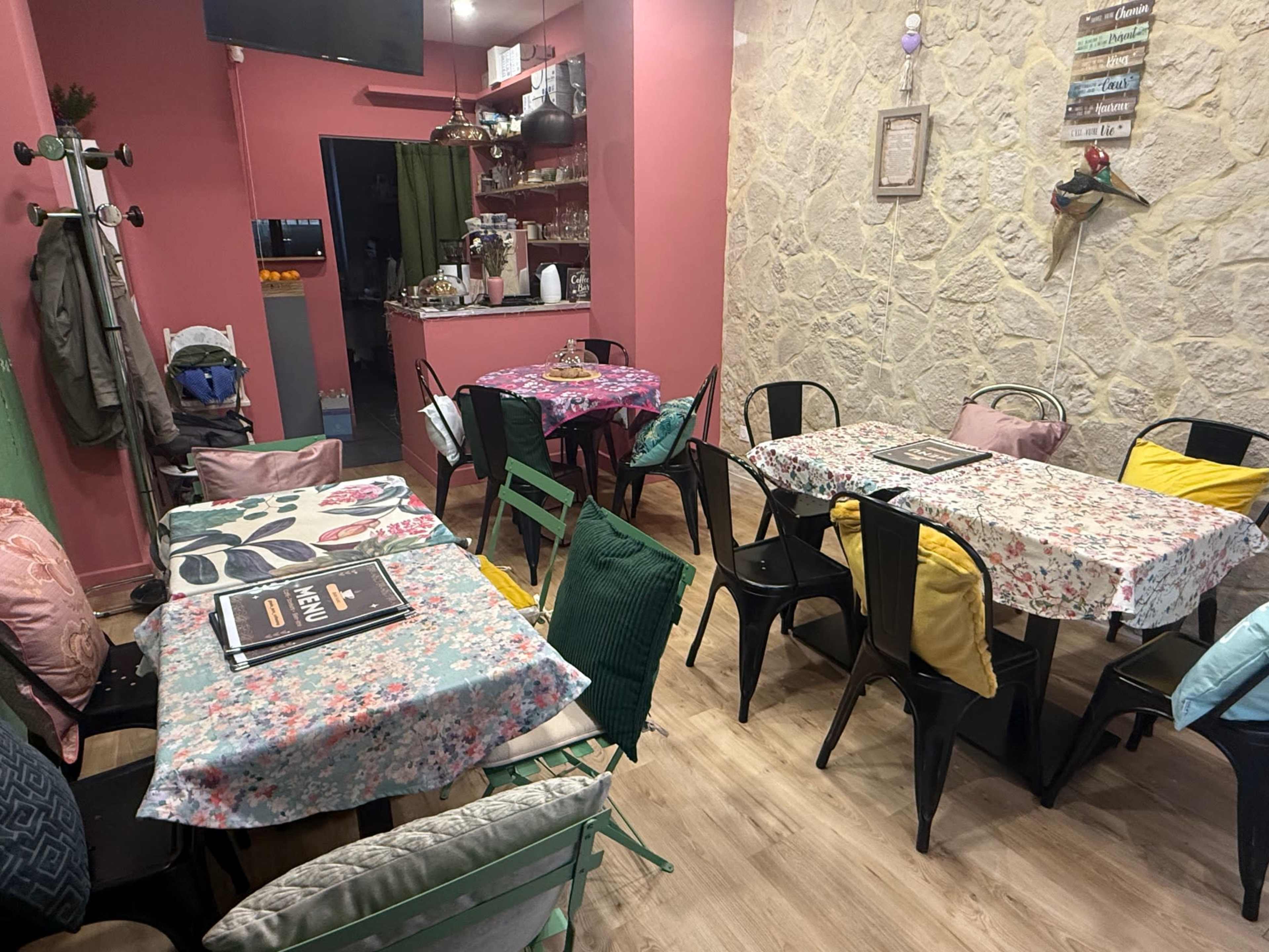 The image shows a brightly colored dining area with multiple tables covered in floral tablecloths and chairs of various styles arranged throughout the space.