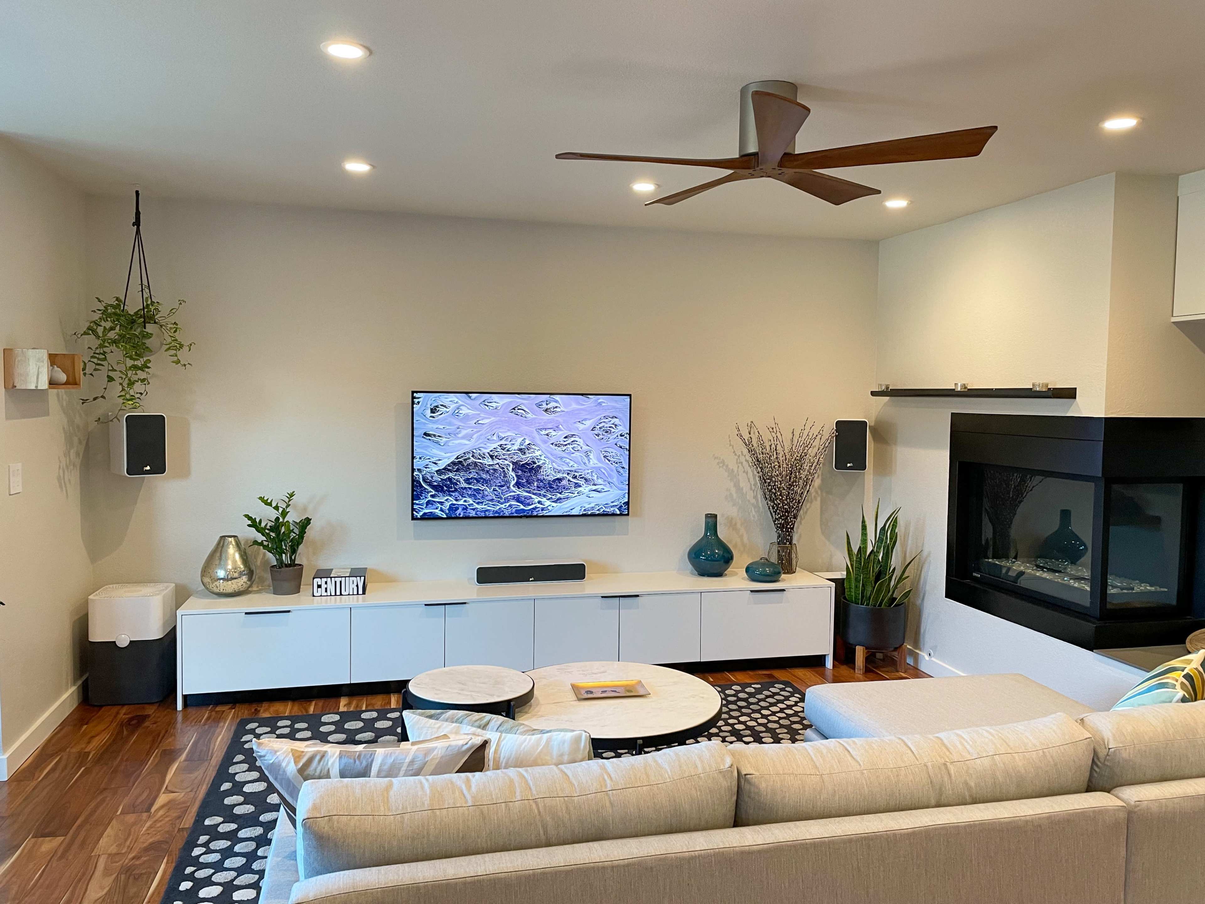 A modern living room with a sectional sofa, a round coffee table, a wall-mounted television, a fireplace, and various decorative plants.
