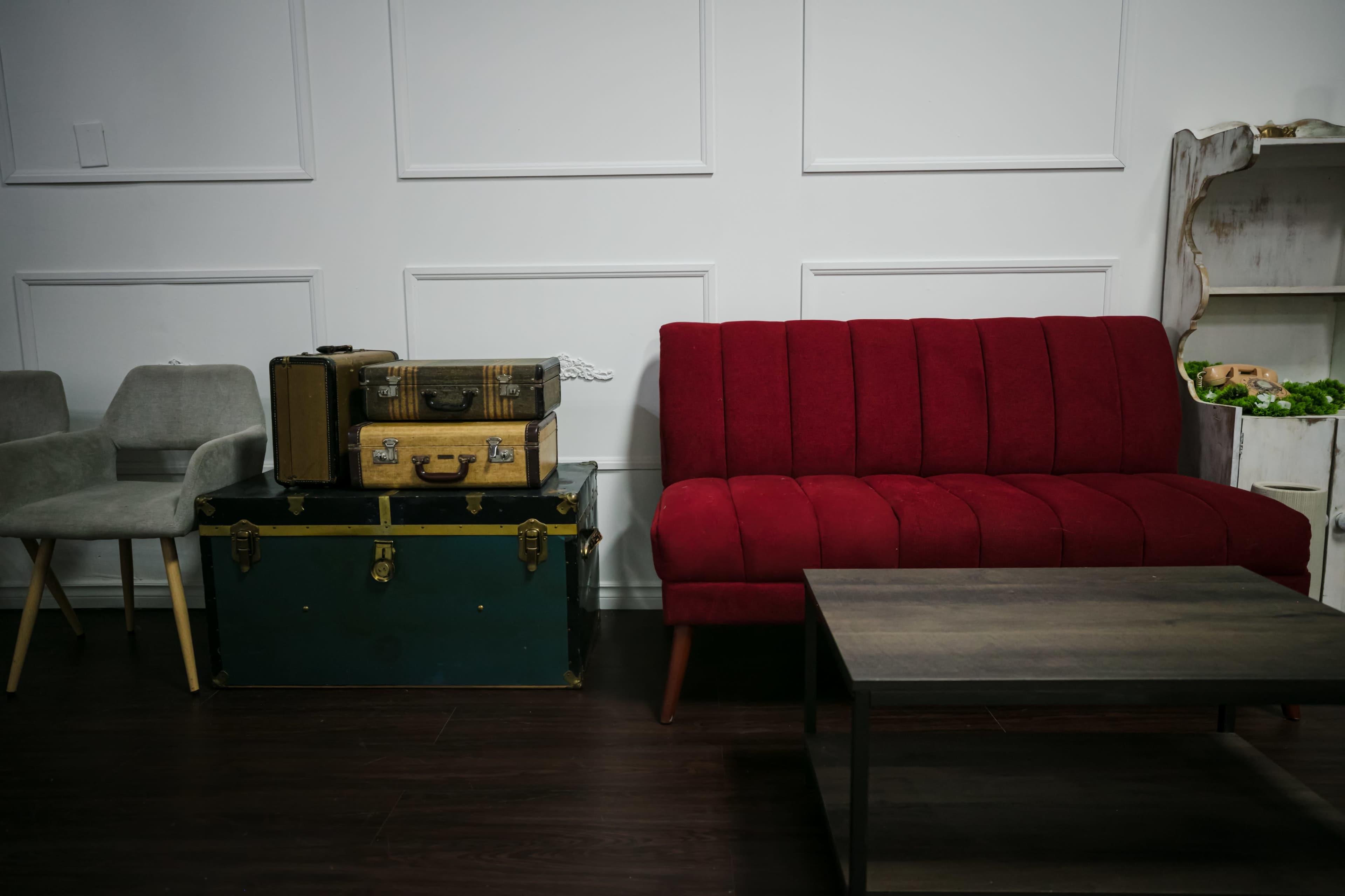A red upholstered sofa is placed next to a wooden coffee table and vintage suitcases against a white paneled wall.