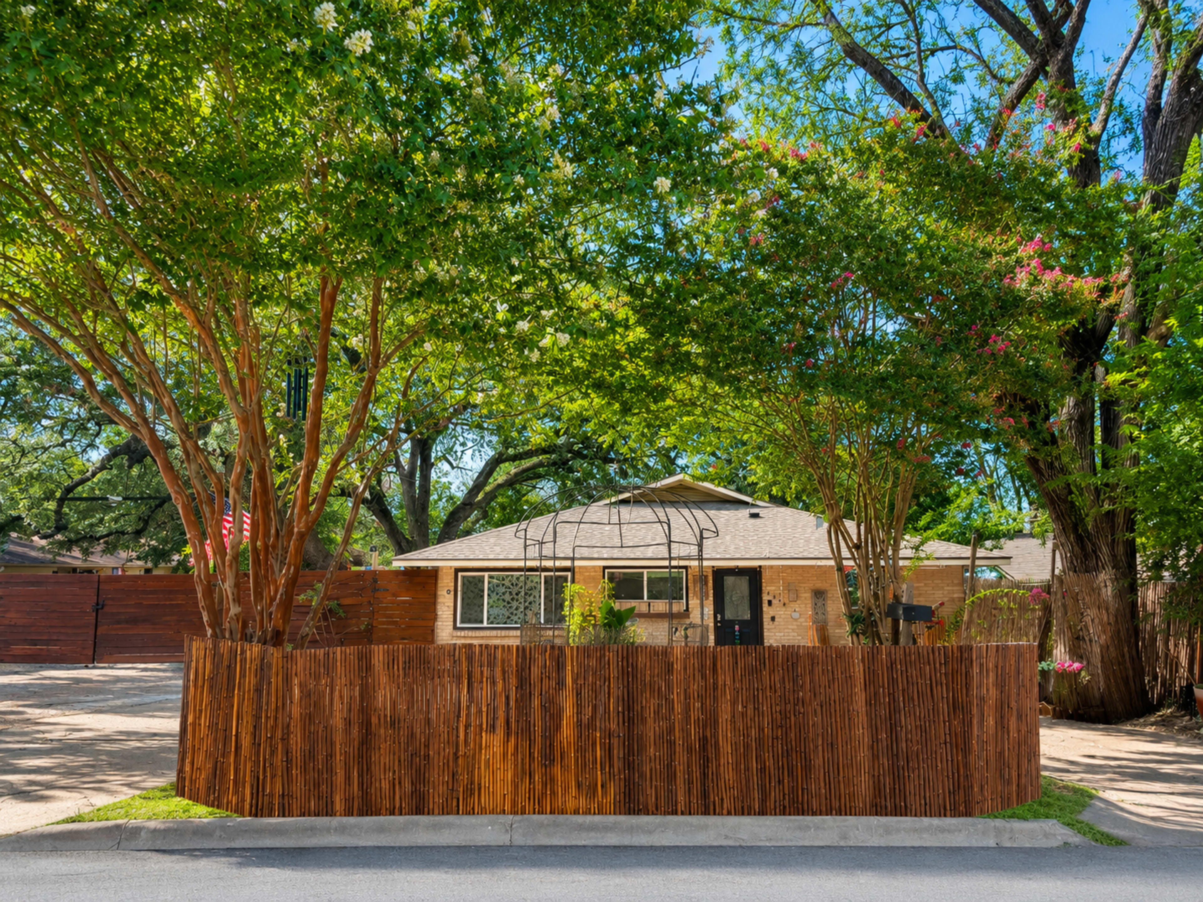 A house with a wooden fence and lush greenery surrounds it, featuring a canopy of trees and vibrant flowers.