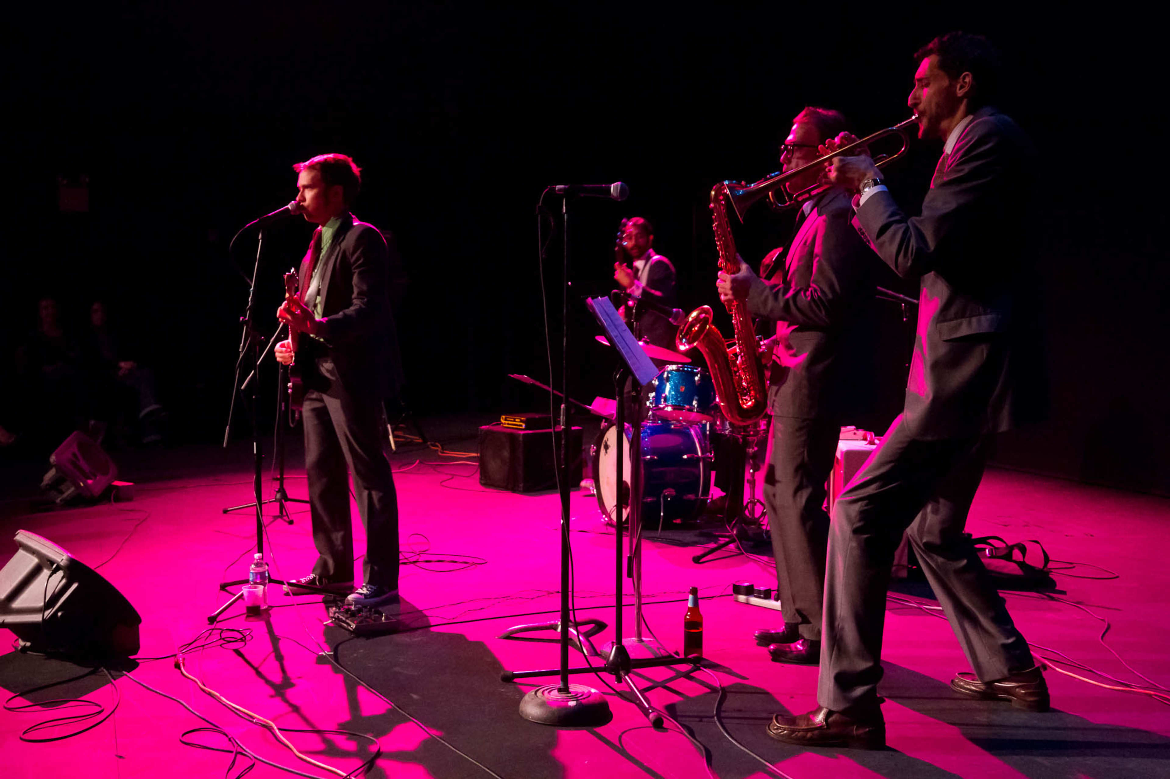 A band performs on stage, with a guitarist and three musicians playing brass instruments, illuminated by pink stage lighting.