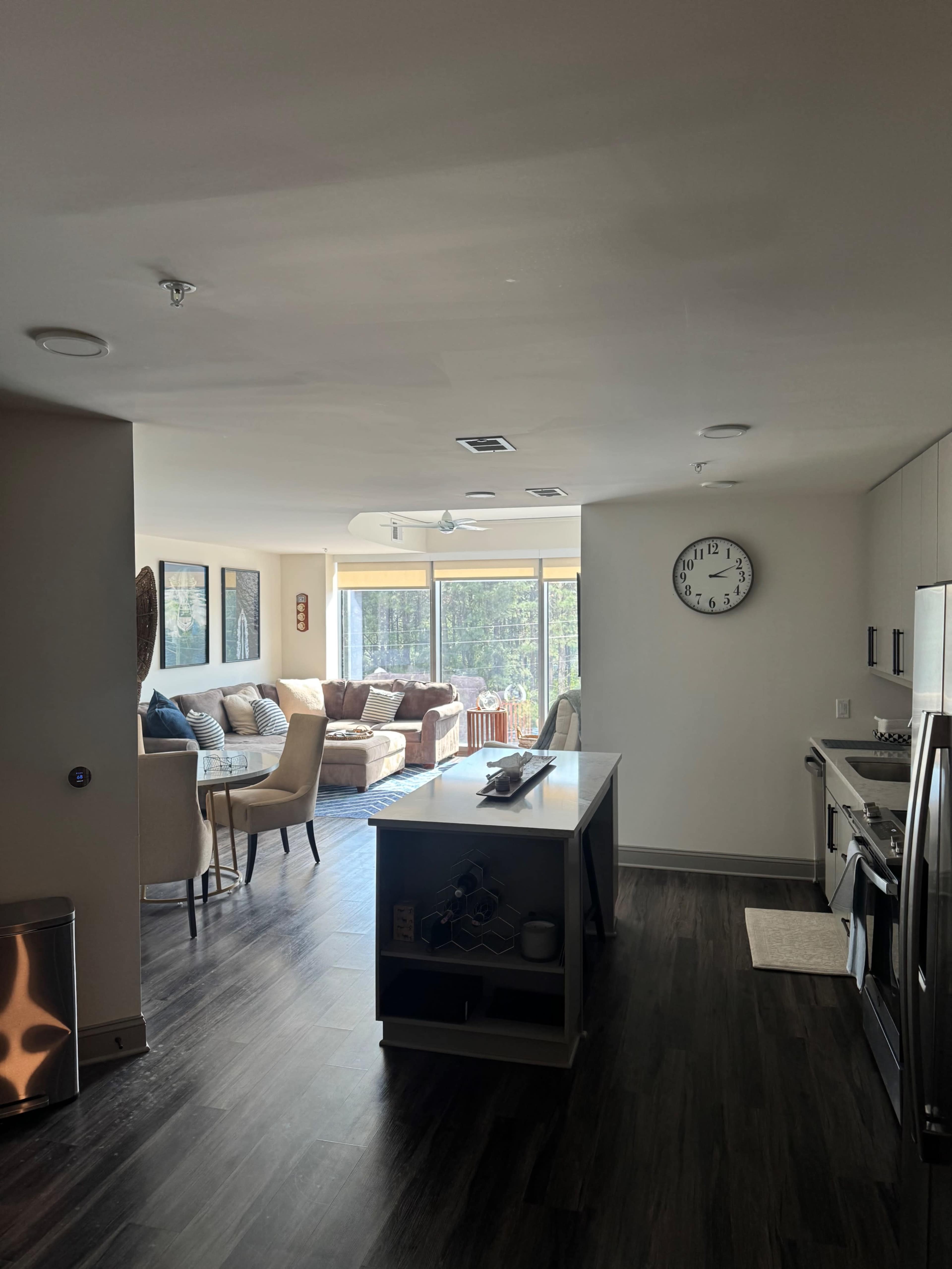 The image shows a modern open-concept living space with a kitchen area on the left and a cozy seating area with large windows on the right.