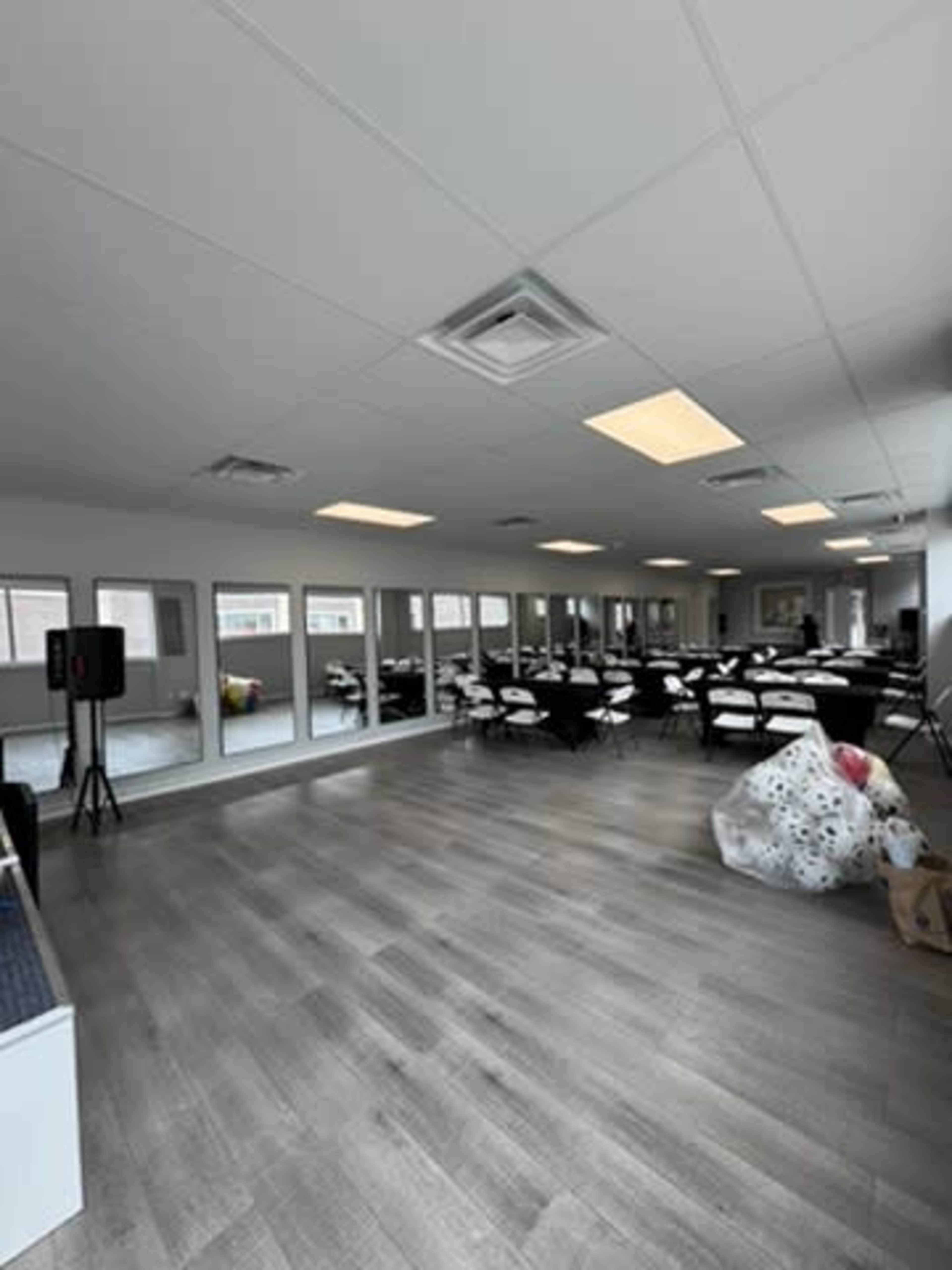 The image shows an indoor event space with several tables and chairs arranged and large windows providing natural light.