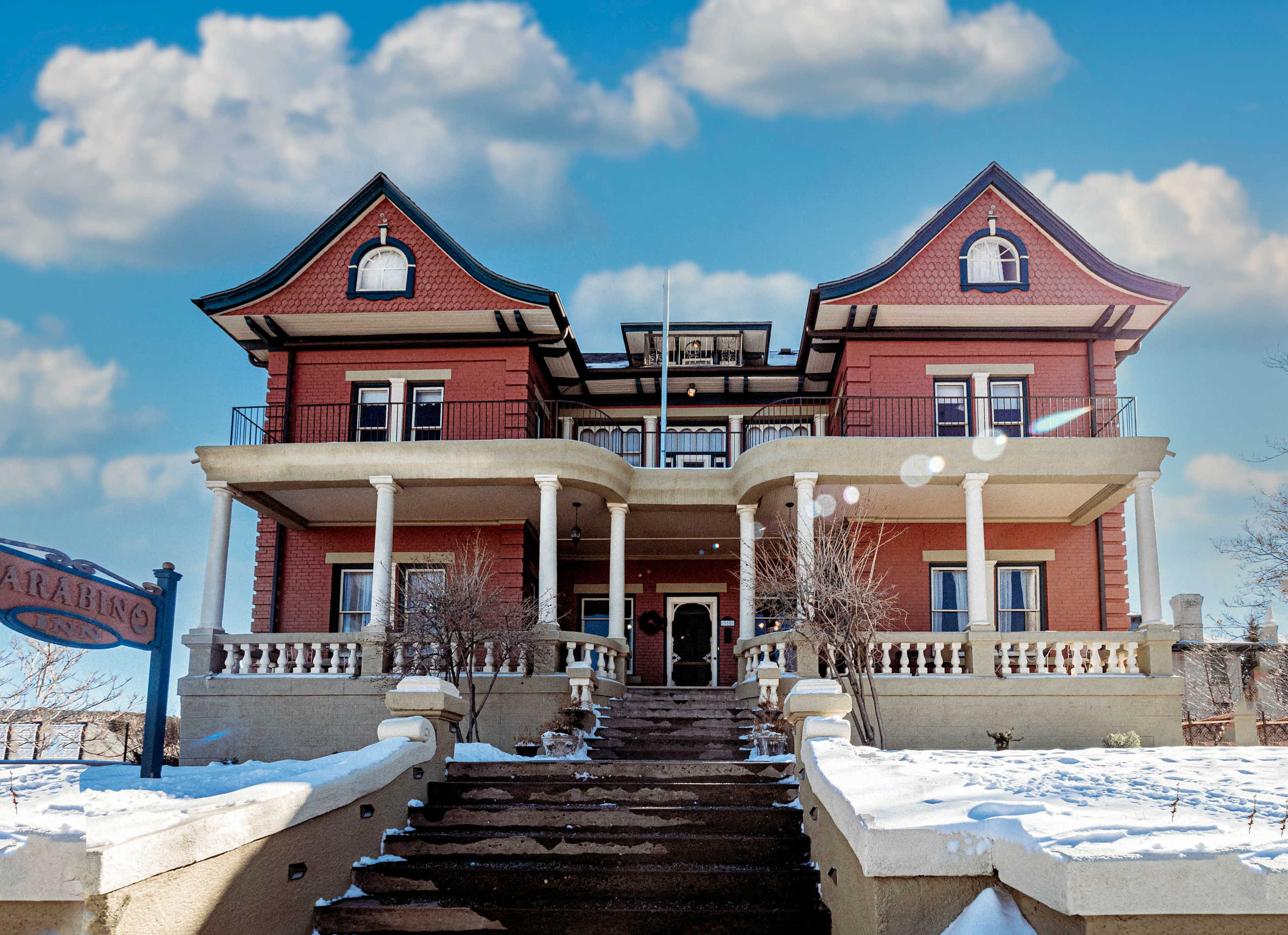 A large, Victorian-style house with a red exterior, multiple balconies, and a staircase leading to the entrance, set against a partly cloudy sky.
