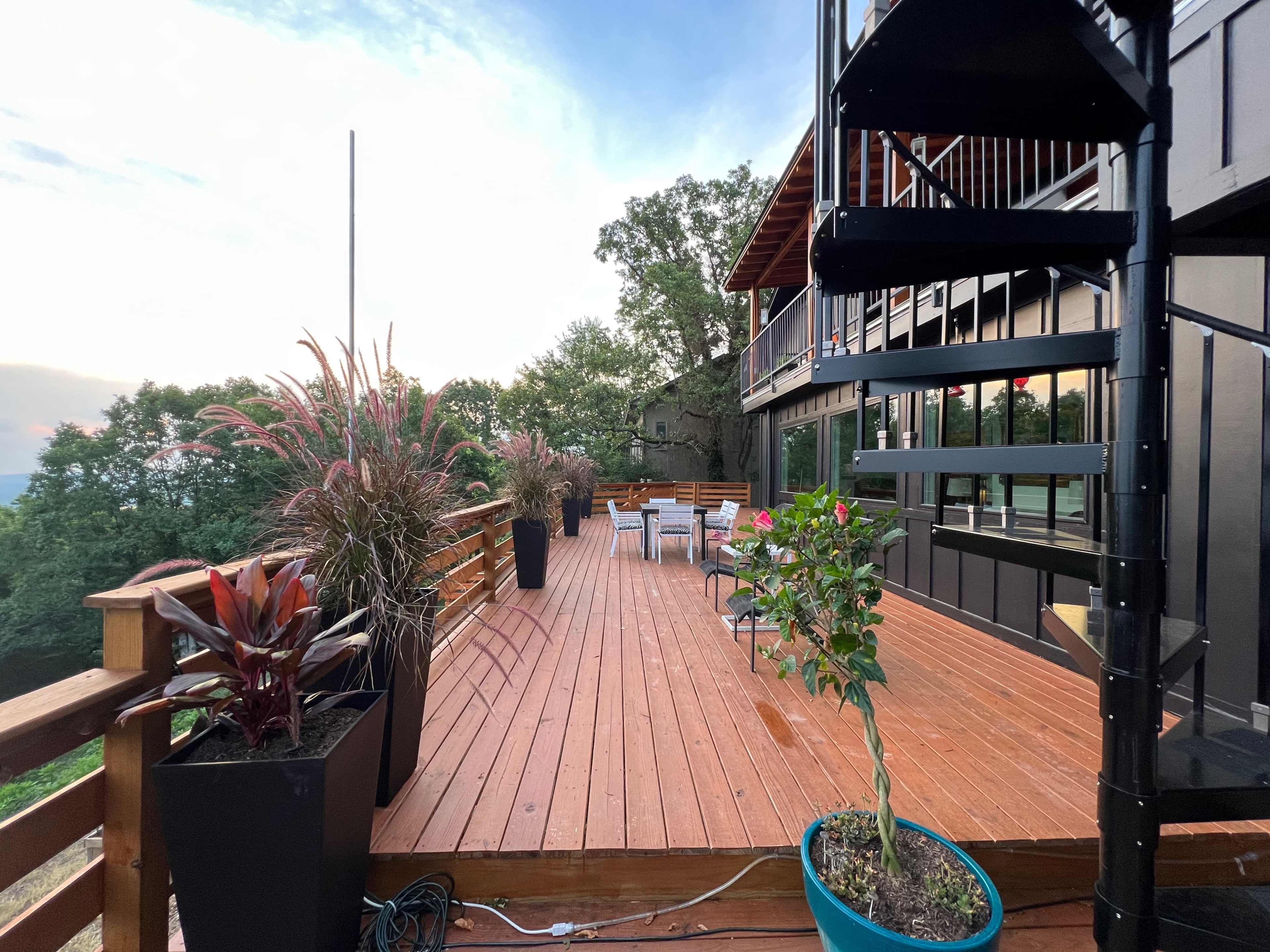 A wooden deck with potted plants and a spiral staircase, overlooking a forested landscape under a cloudy sky.